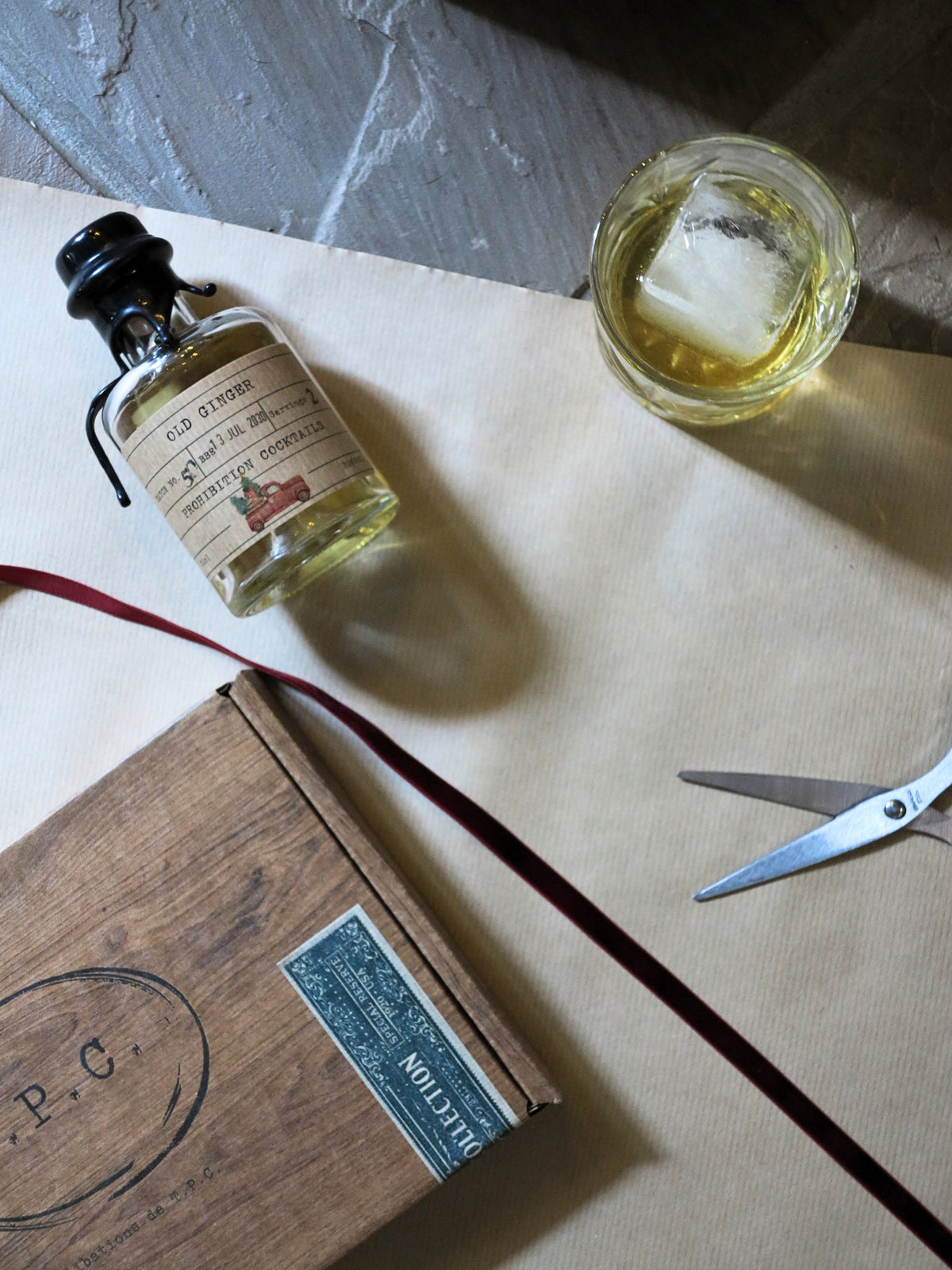 A styled flat-lay featuring a miniature bottle of whiskey with a vintage-style label, a glass of whiskey with an ice cube, a wooden box with a blue label, and silver scissors on a beige paper background.