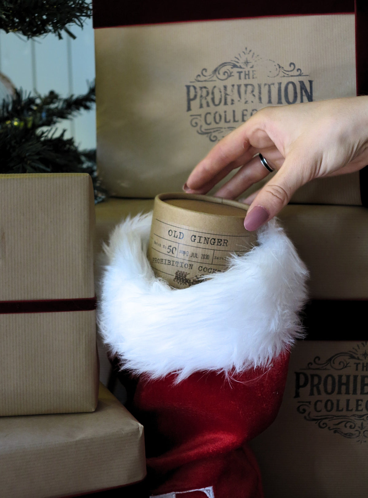 A hand with pink nail polish holds a small cylindrical tin labeled 'OLD GINGER' above a red and white faux-fur-trimmed stocking. The scene includes wrapped gift boxes and paper bags printed with 'The Prohibition Collection' in the background.