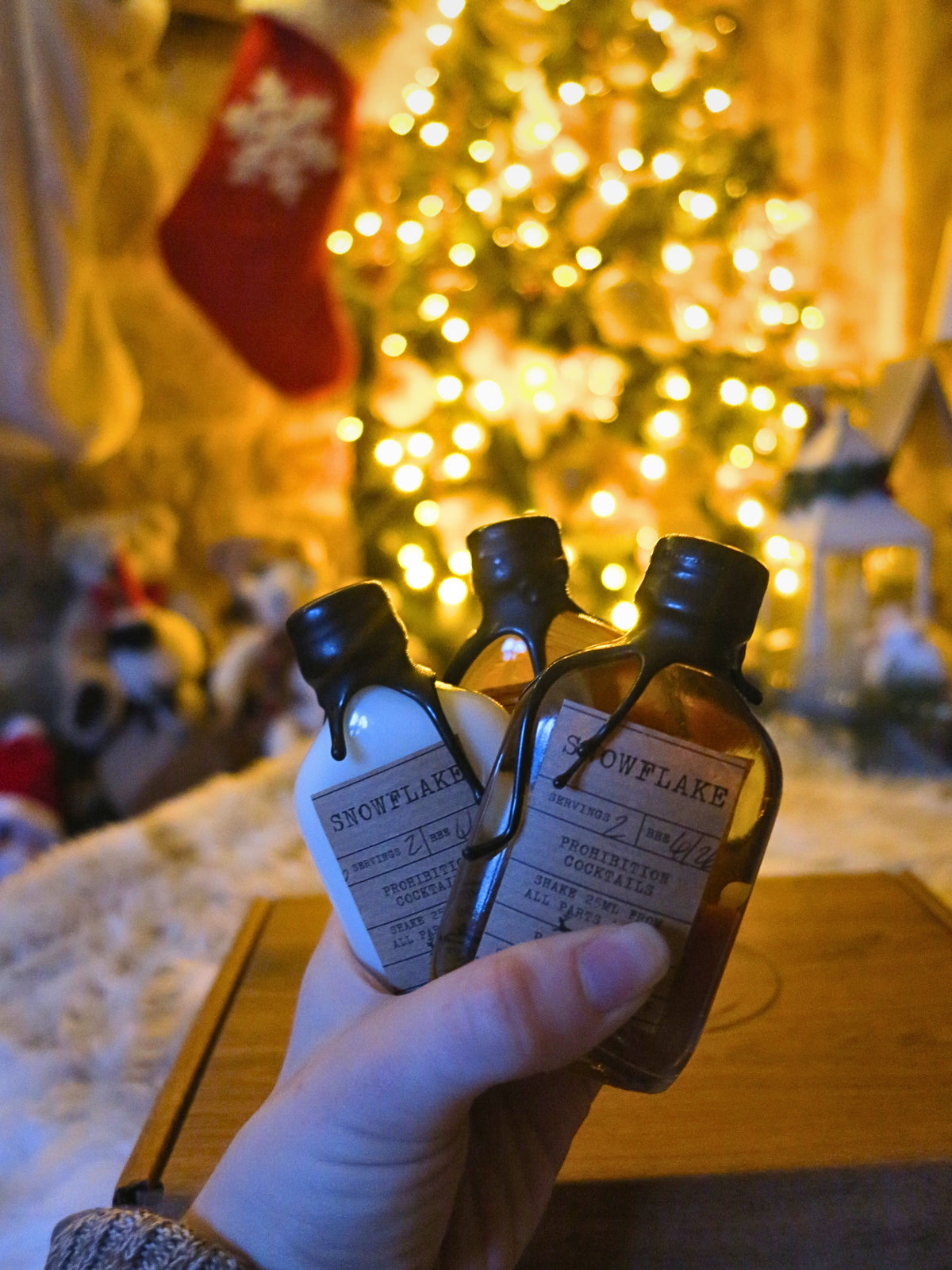A hand holds three miniature decorative bottles labeled 'SNOWFLAKE' in front of a softly blurred Christmas tree with warm lights and a red stocking in the background.
