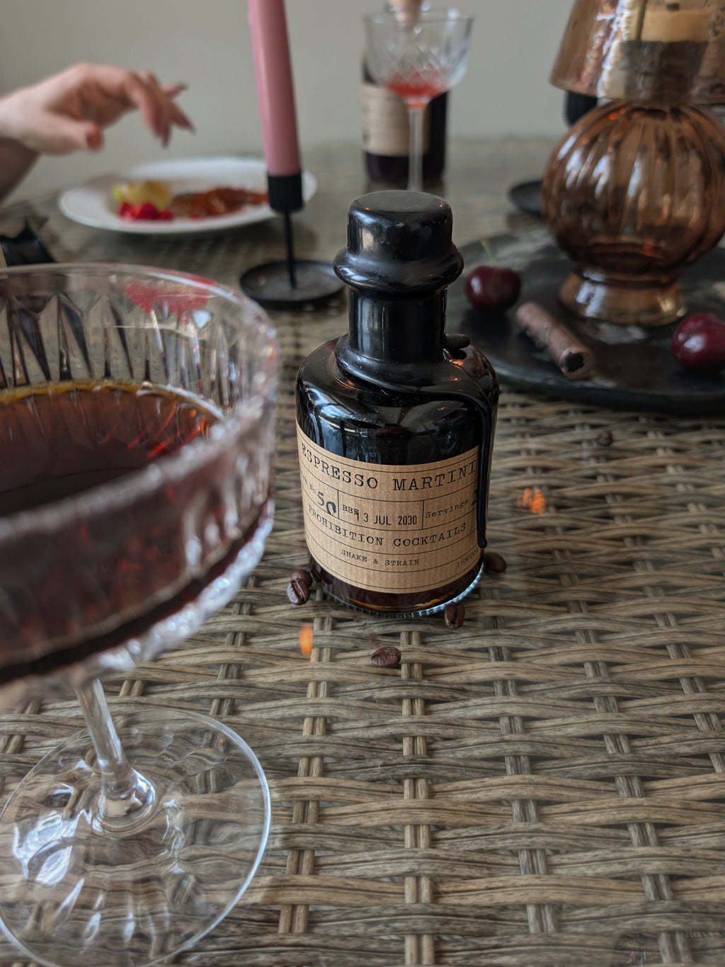 A small dark glass bottle labeled 'ESPRESSO MARTINI' sits on a woven table mat beside a stemmed glass filled with dark red liquid, with candles, cherries, and other bar-like props in a softly blurred indoor setting.