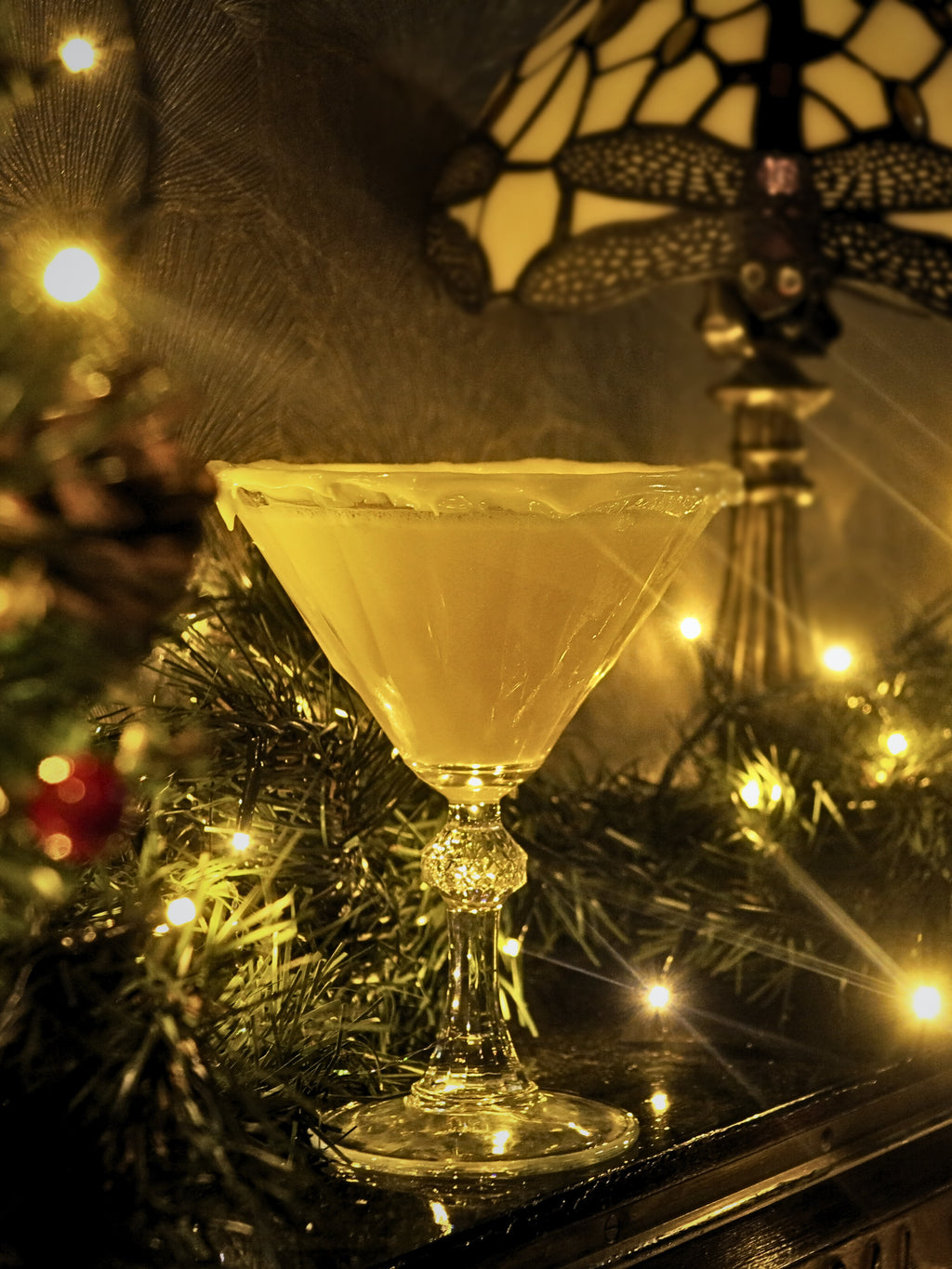 A clear martini-style coupe glass filled with a pale yellow drink, set on a dark tabletop with holiday greenery, warm string lights, and a stained-glass-style lamp in the background.