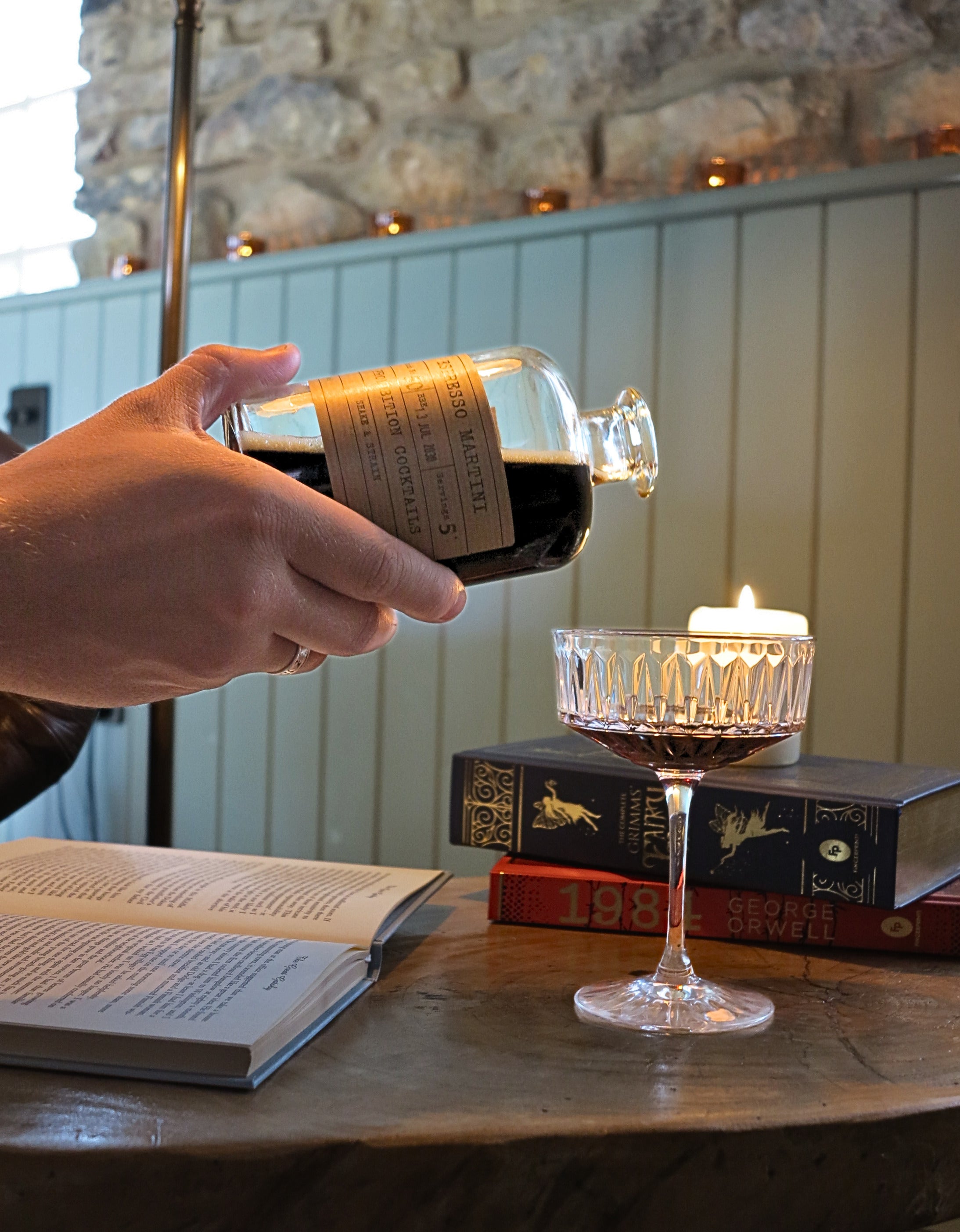 A hand pours a dark liquid from a glass bottle with a cream label onto a crystal cocktail coupe glass resting on stacked books, with an open book and a lit candle in a rustic indoor setting. The label reads: Espresso Martini by Prohibition Cocktails