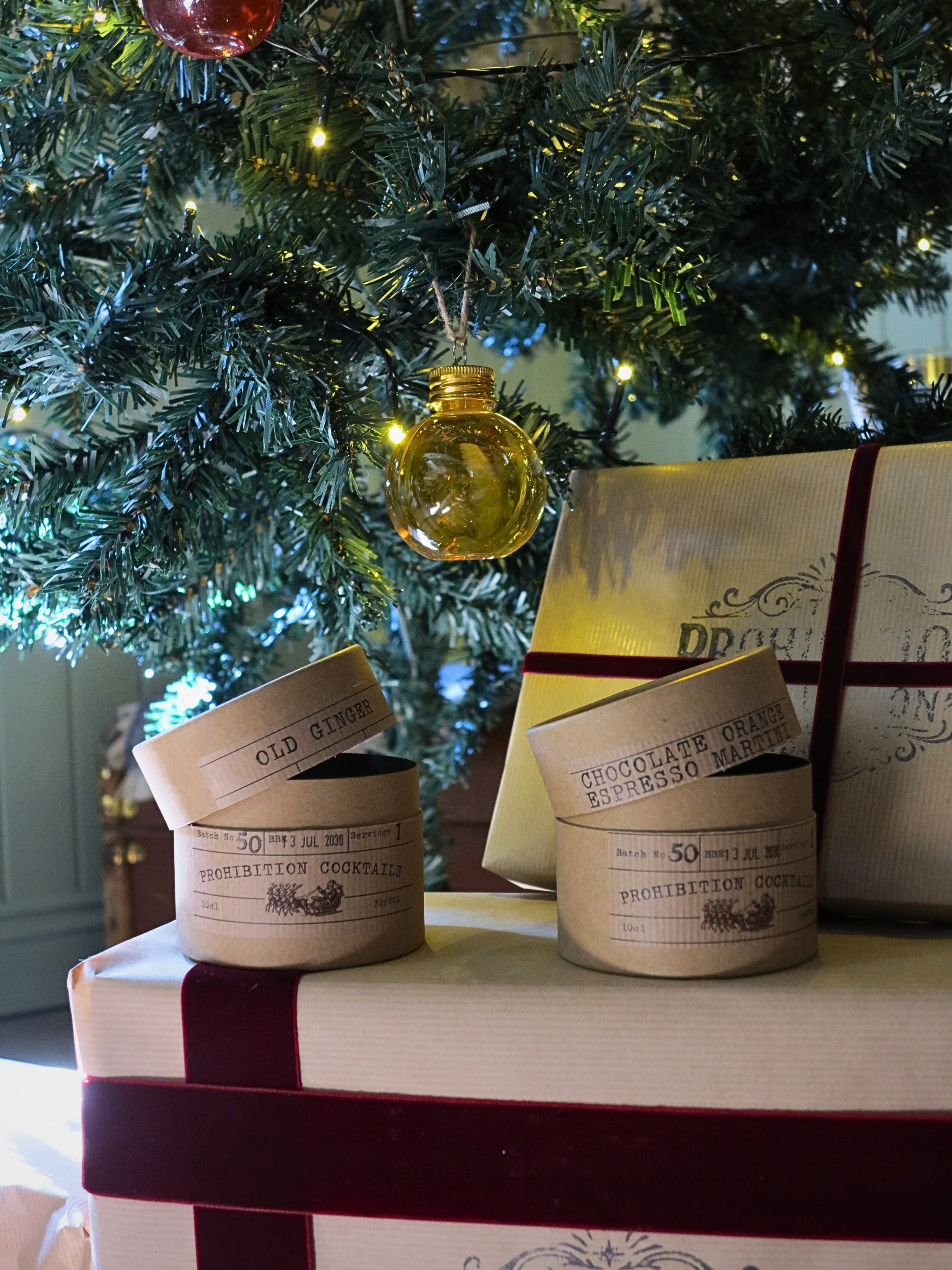 Two gift boxes with lids off, labeled 'Old Ginger' and 'Chocolate Orange Espresso Martini', arranged on top of a wrapped gift box under a decorated Christmas tree with lights and ornaments.