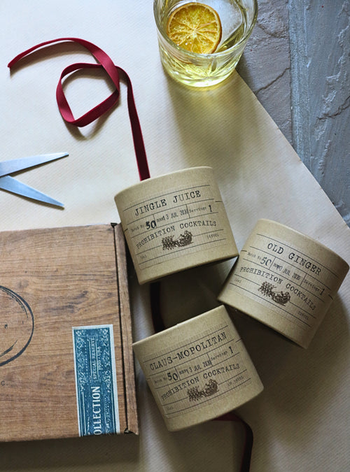 Four decorative paper-wrapped cocktail mixers from Prohibition Cocktails are arranged on a beige paper background with a wooden box, scissors, a red ribbon, and a glass of tea with a lemon slice. The labels show flavors: Jingle Juice, Old Ginger, and Claus-mopolitan