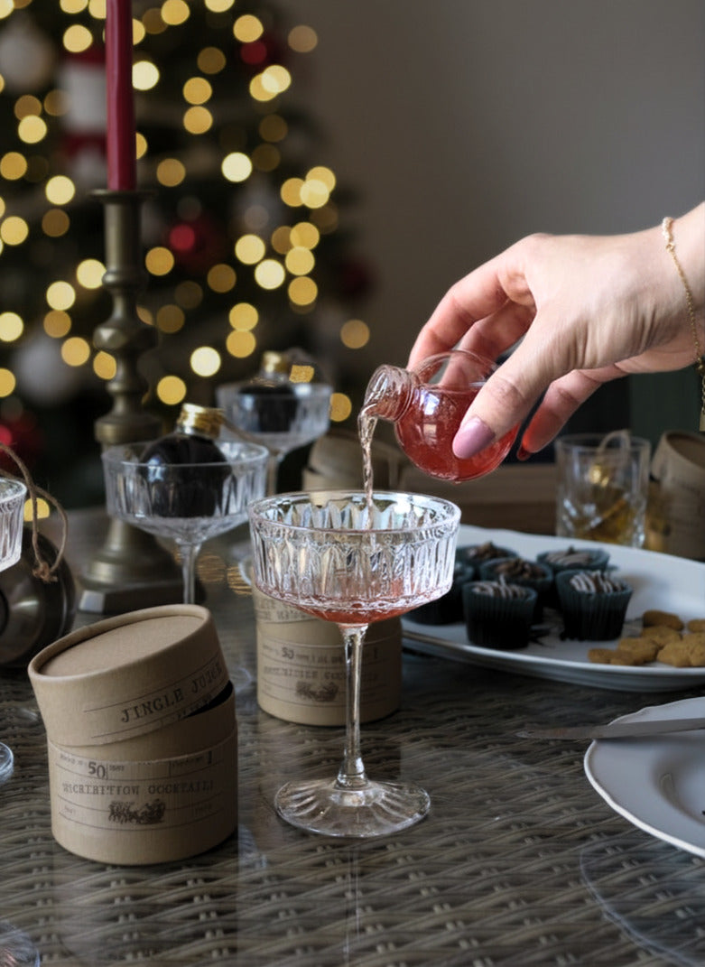 A hand pours a pink beverage - jingle juice - from a small glass pitcher into a clear cut-glass coupe coupe-style glass on a dining table, with candles, plates, and blurred holiday lights in the background.