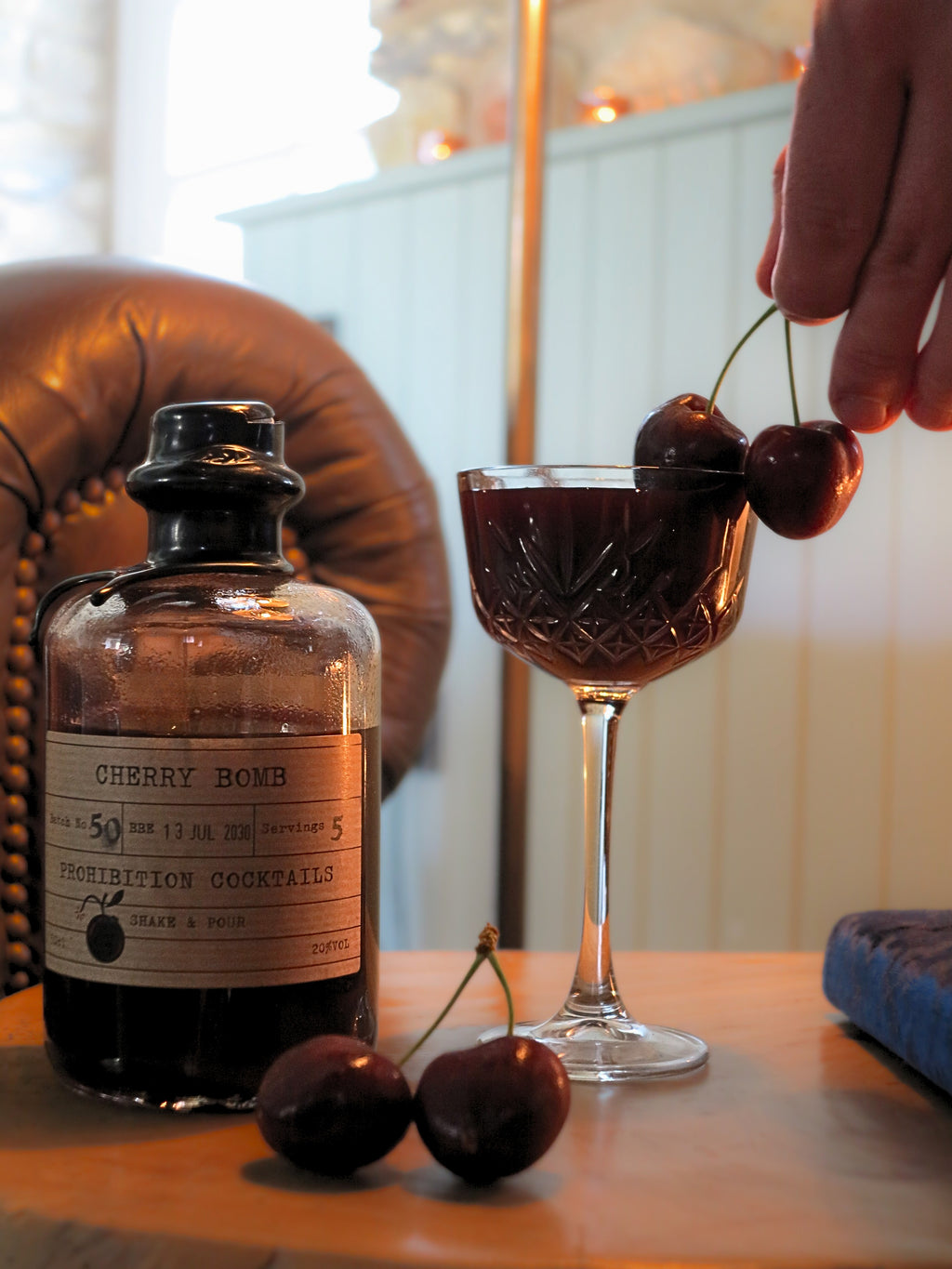A bottle labeled Cherry Bomb Prohibition Cocktails sits on a wooden table beside a coupe glass filled with a dark red cocktail, with cherries on the glass rim and two cherries on the table. A hand is placing a cherry onto the glass, and a leather chair and indoor setting are visible in the background.