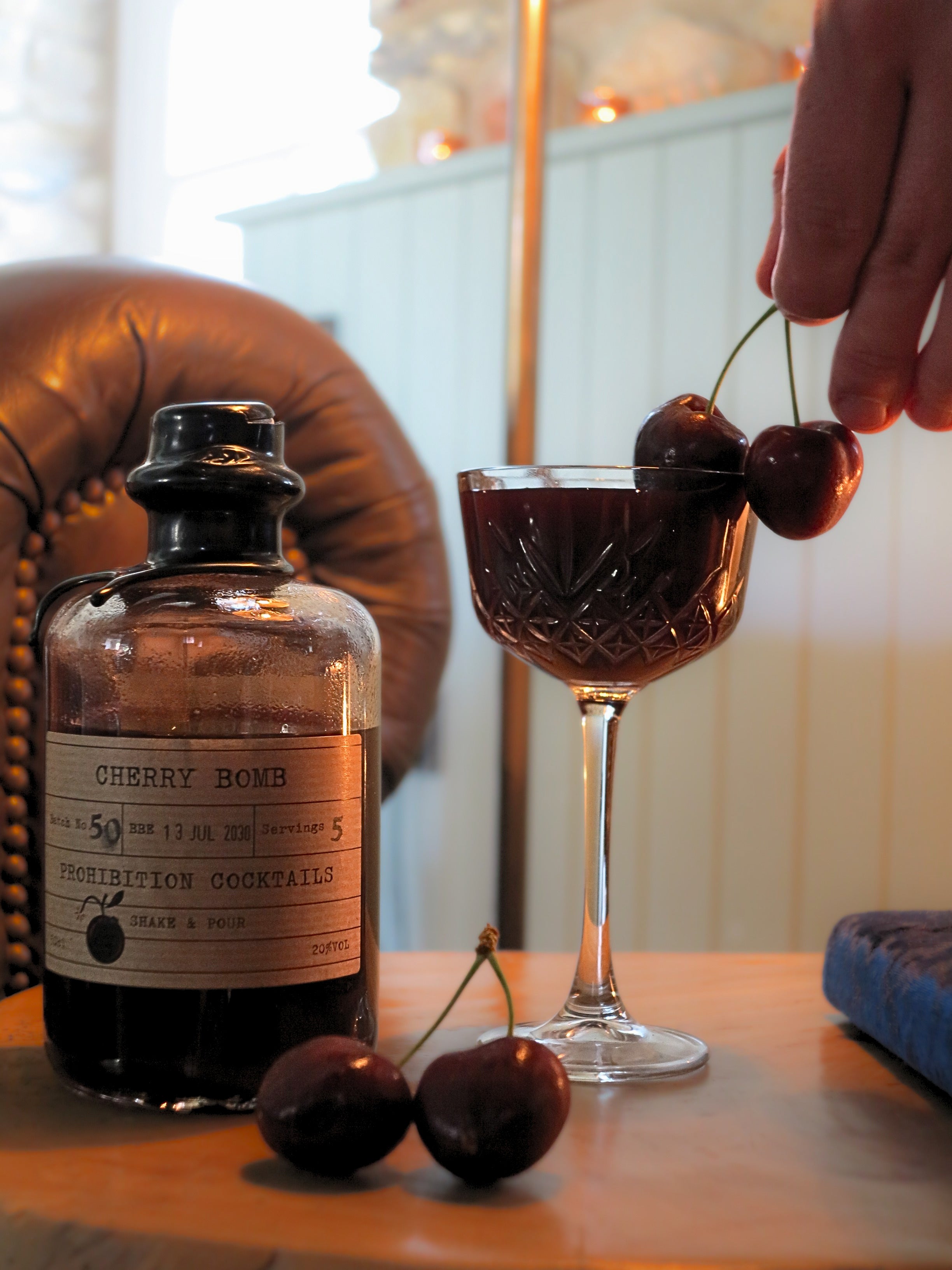 A bottle labeled Cherry Bomb Prohibition Cocktails sits on a wooden table beside a coupe glass filled with a dark red cocktail, with cherries on the glass rim and two cherries on the table. A hand is placing a cherry onto the glass, and a leather chair and indoor setting are visible in the background.