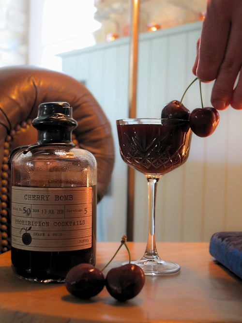 A bottle labeled Cherry Bomb Prohibition Cocktails sits on a wooden table beside a coupe glass filled with a dark red cocktail, with cherries on the glass rim and two cherries on the table. A hand is placing a cherry onto the glass, and a leather chair and indoor setting are visible in the background.