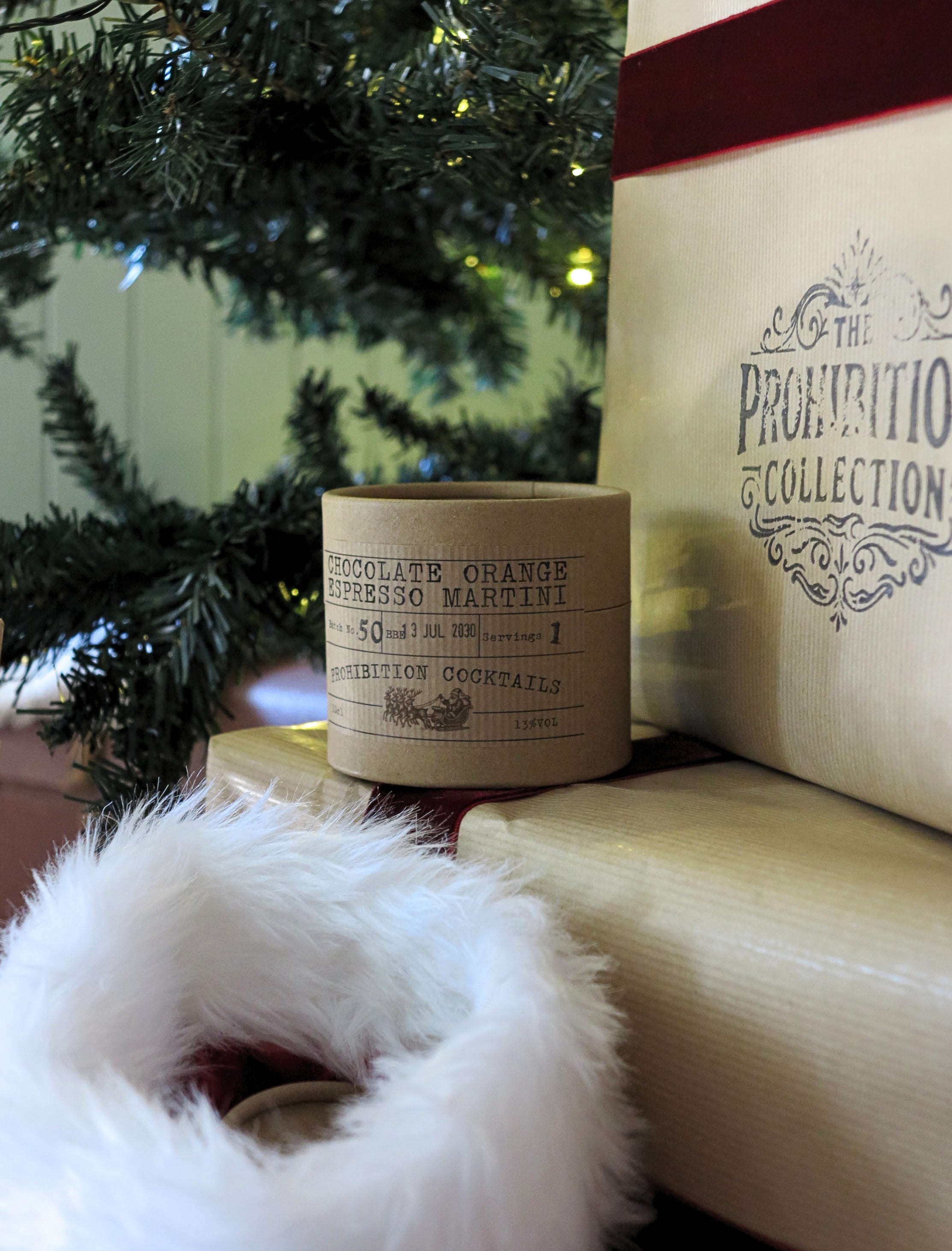 A close-up of a wrapped gift featuring a cream-colored paper sleeve with black text reading 'CHOCOLATE ORANGE ESPRESSO MARTINI' A larger cream gift bag from 'The Prohibition Collection' is visible behind it, with a white faux-fur trim in the foreground and a Christmas tree in the background.