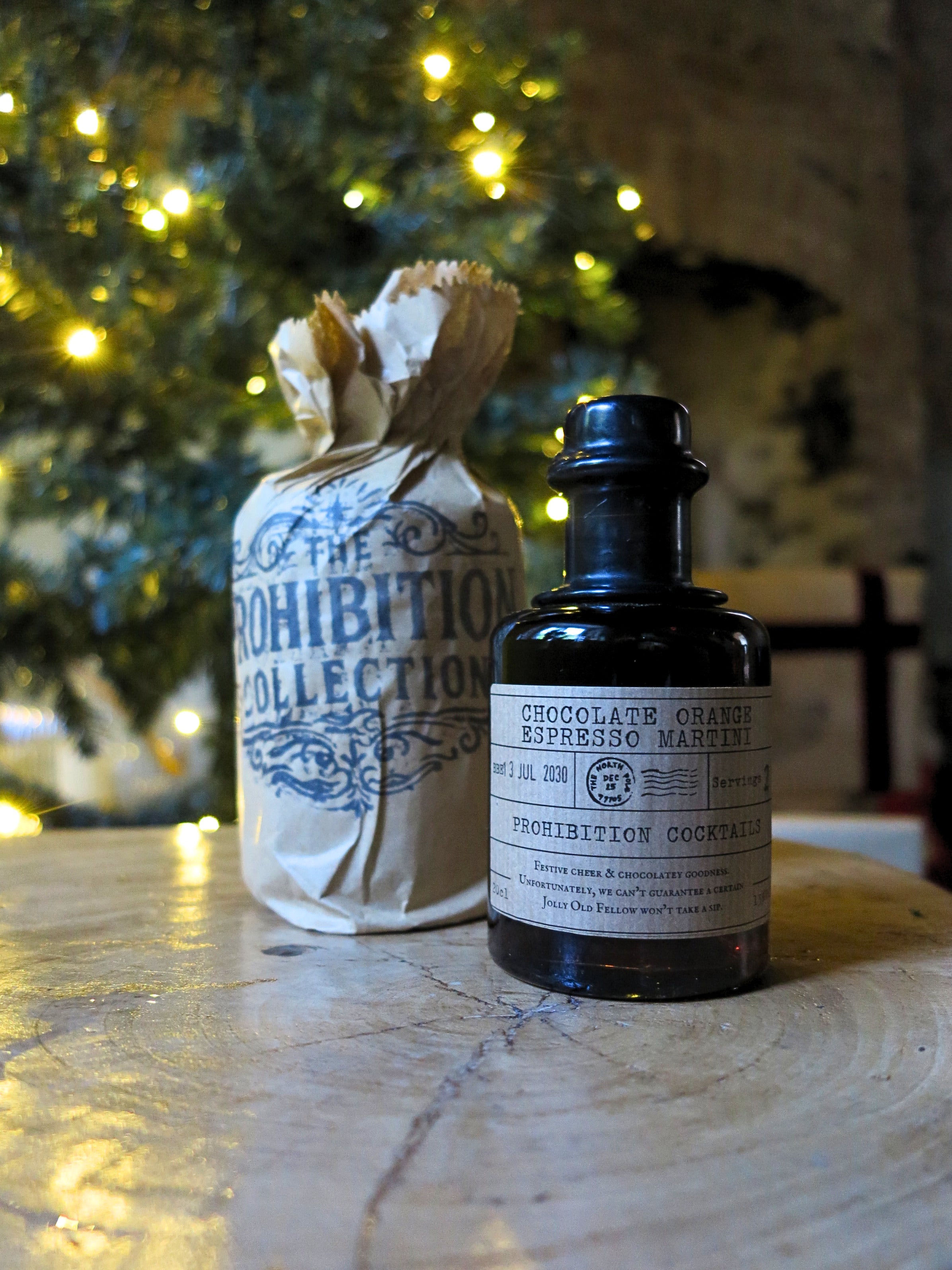 A dark glass bottle labeled 'Chocolate Orange Espresso Martini' stands on a stone surface next to a paper bag printed with 'The Prohibition Collection' and tied with twine, with a blurred festive tree of lights in the background.