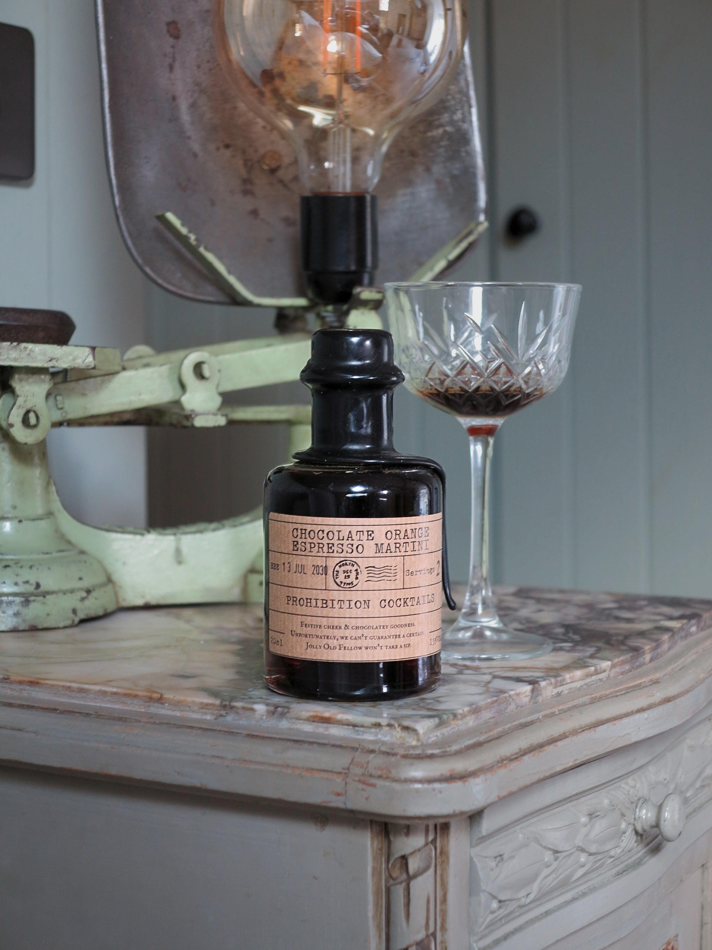 A dark glass bottle of chocolate orange espresso martini by Prohibition Cocktails with a vintage paper label sits on a distressed wooden table next to a cut-glass coupe glass and a large antique lamp.