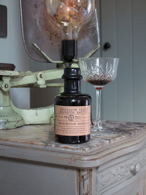 A dark glass bottle of chocolate orange espresso martini by Prohibition Cocktails with a vintage paper label sits on a distressed wooden table next to a cut-glass coupe glass and a large antique lamp.