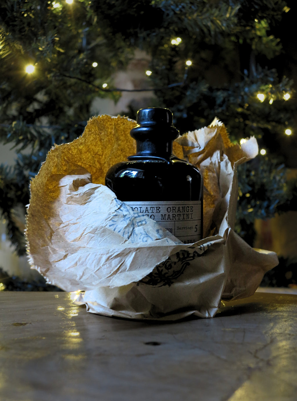 A dark glass bottle with a black stopper and a partially visible label reading 'chocolate orange espresso martini' sits wrapped in crumpled white paper with a gold textured accent, placed on a tabletop in front of a softly blurred Christmas tree with warm lights.
