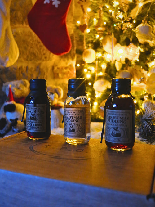 Three small bottles of Christmas-themed liqueurs from The Prohibition Collection are lined up on a wooden surface in front of a warm, glowing Christmas tree and stockings.