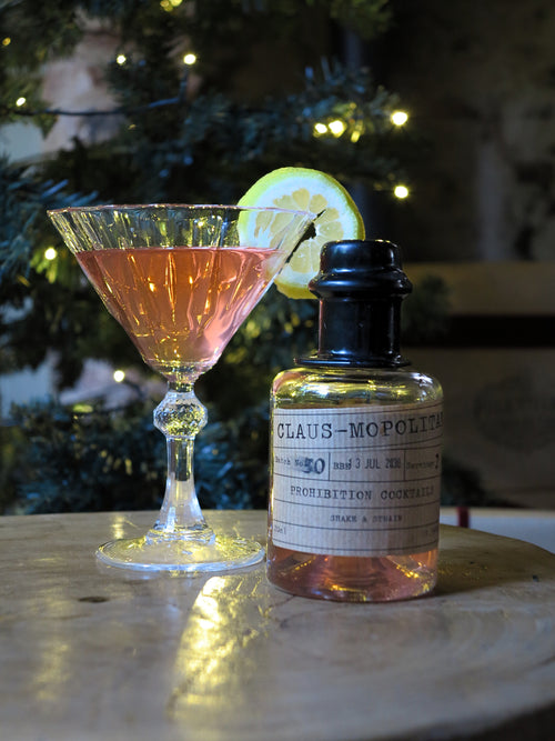 A pink prohibition cocktail in a coupe glass with a lemon wheel garnish, placed beside a bottle labeled 'Claus-Mopolitan Prohibition Cocktails' on a wooden surface with blurred holiday lights in the background.