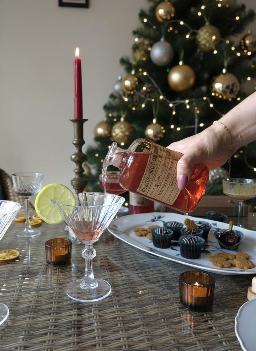 A hand pours a pinkish-red liquid from a bottle into a coupe glass on a decorated holiday table with a Christmas tree in the background.