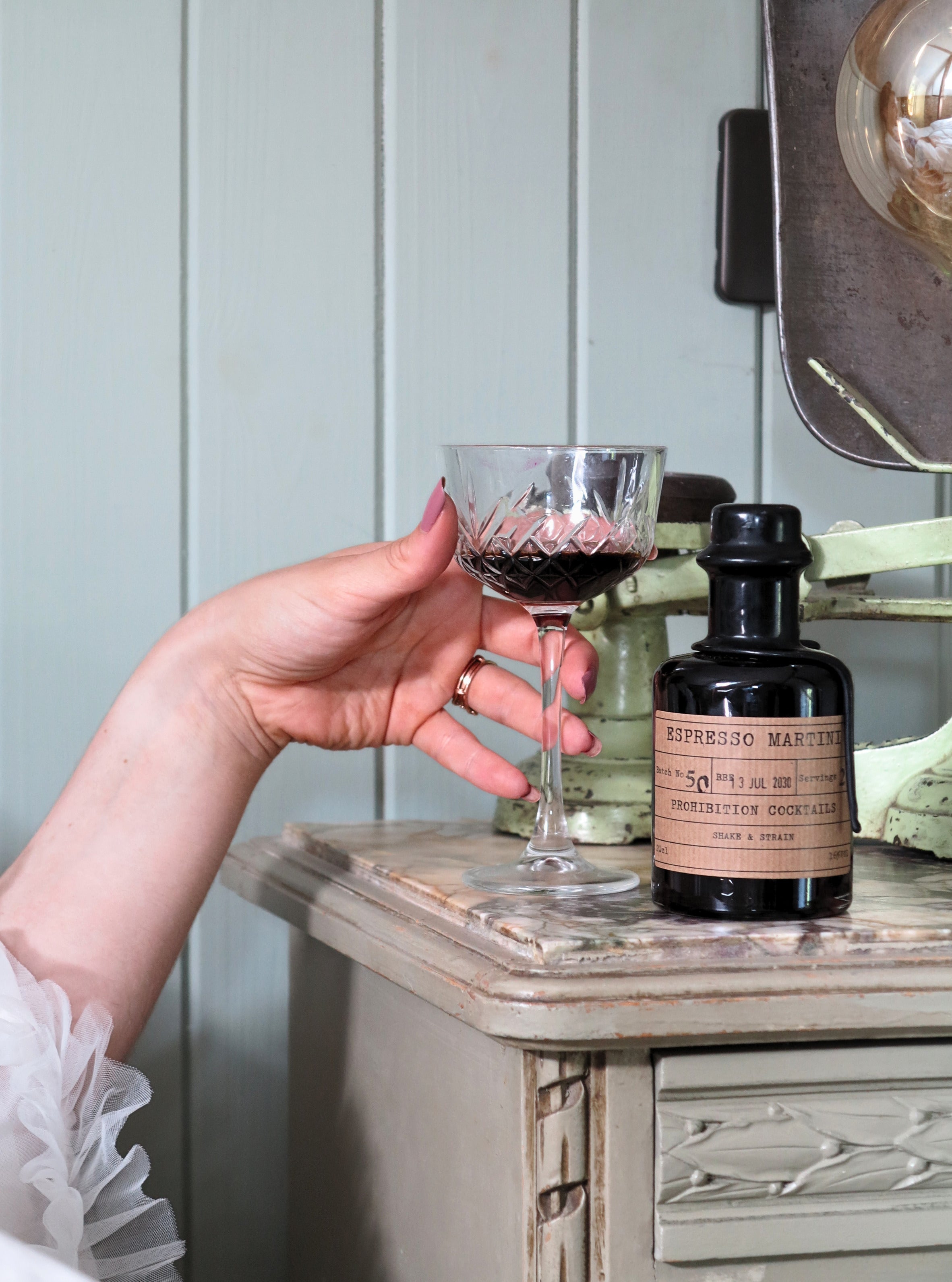 A hand holds a coupe glass containing a dark liquid next to a bottle of Espresso Martini mix by Prohibition Cocktails on a distressed wooden table, with a light paneled wall in the background.
