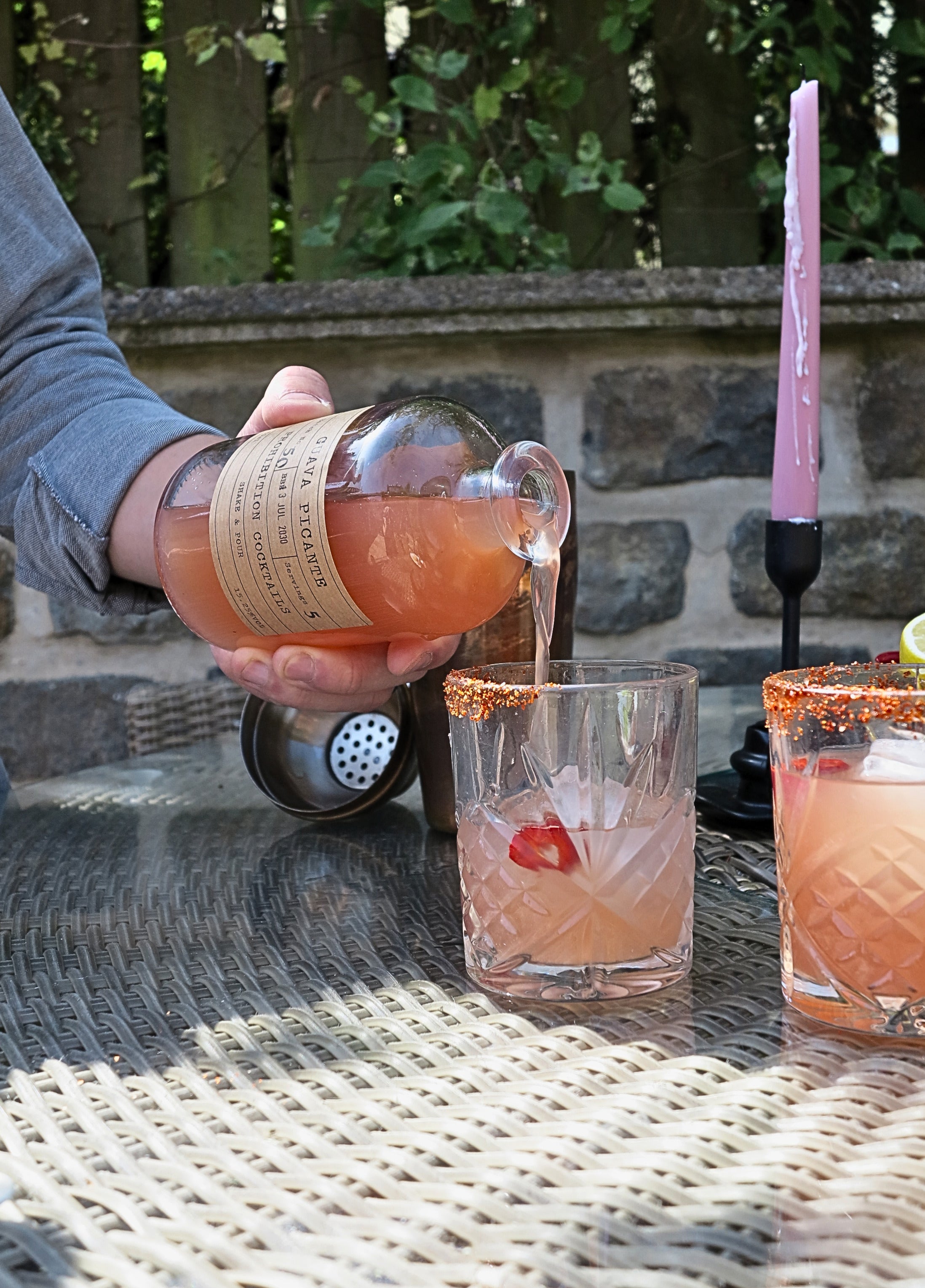 A person pours a guava picante from Prohibition Cocktails from a glass bottle into a clear rocks glass with a sugar rim, set on an outdoor patio table.
