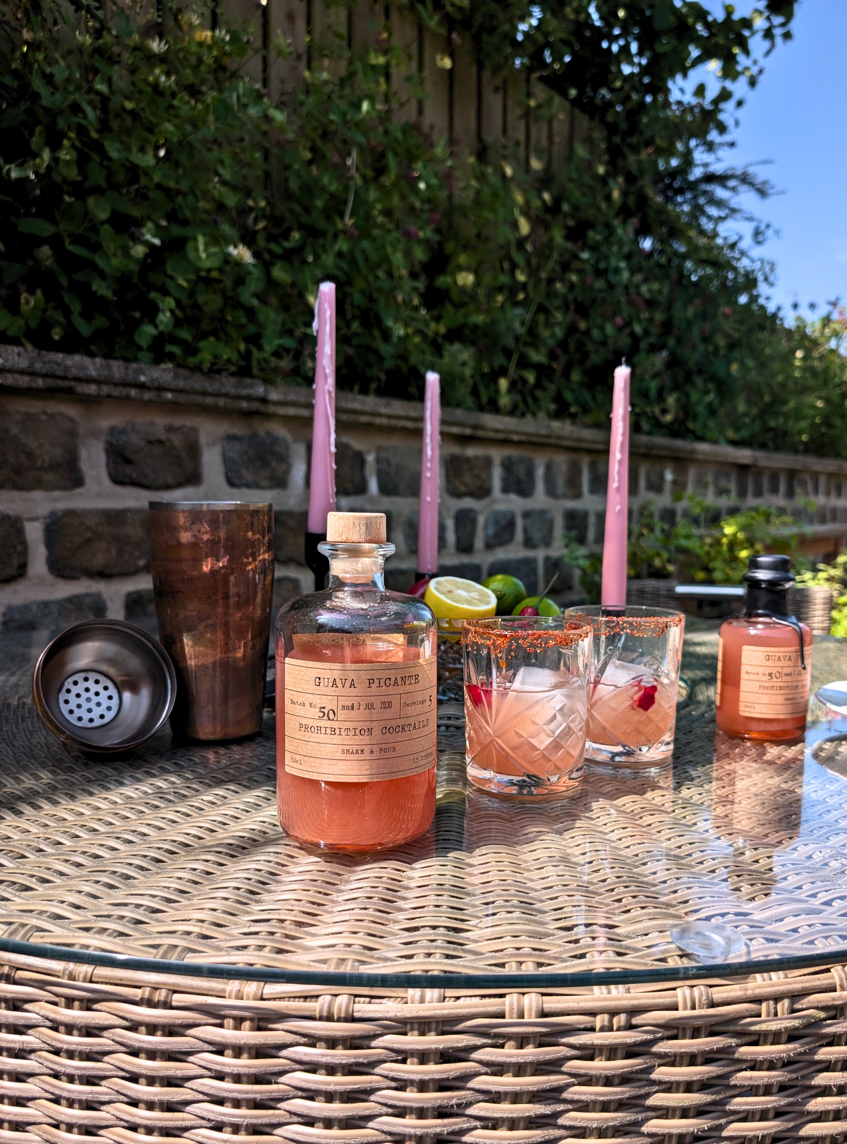 A bottle of guava picante by prohibition cocktails and two matching glasses with pink straws sit on a wicker table outdoors, alongside a copper shaker and lime slices, with a stone wall and greenery in the background.