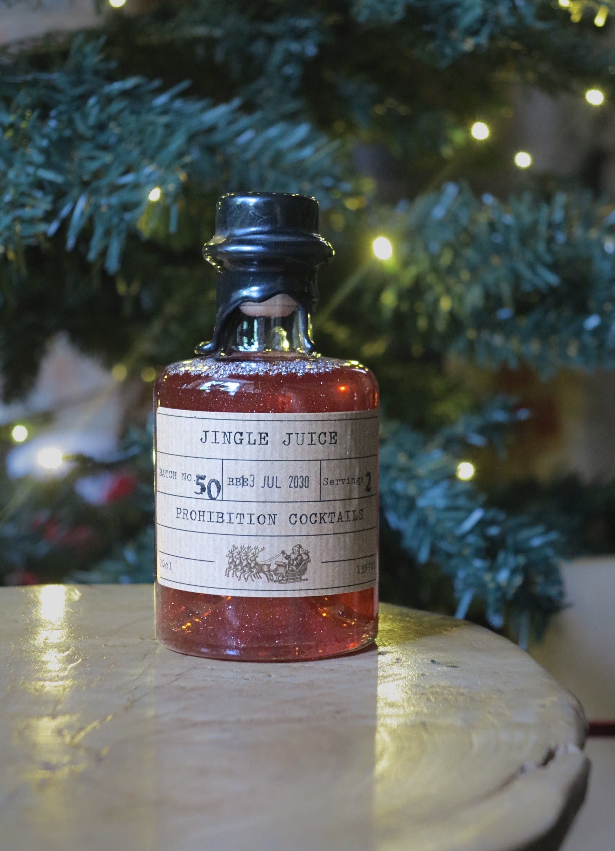 A small glass bottle filled with amber liquid sits on a light wooden surface, with a blurred Christmas tree and warm string lights in the background. The bottle has a black cap and a vintage-style paper label reading 'JINGLE JUICE' and 'PROHIBITION COCKTAILS.