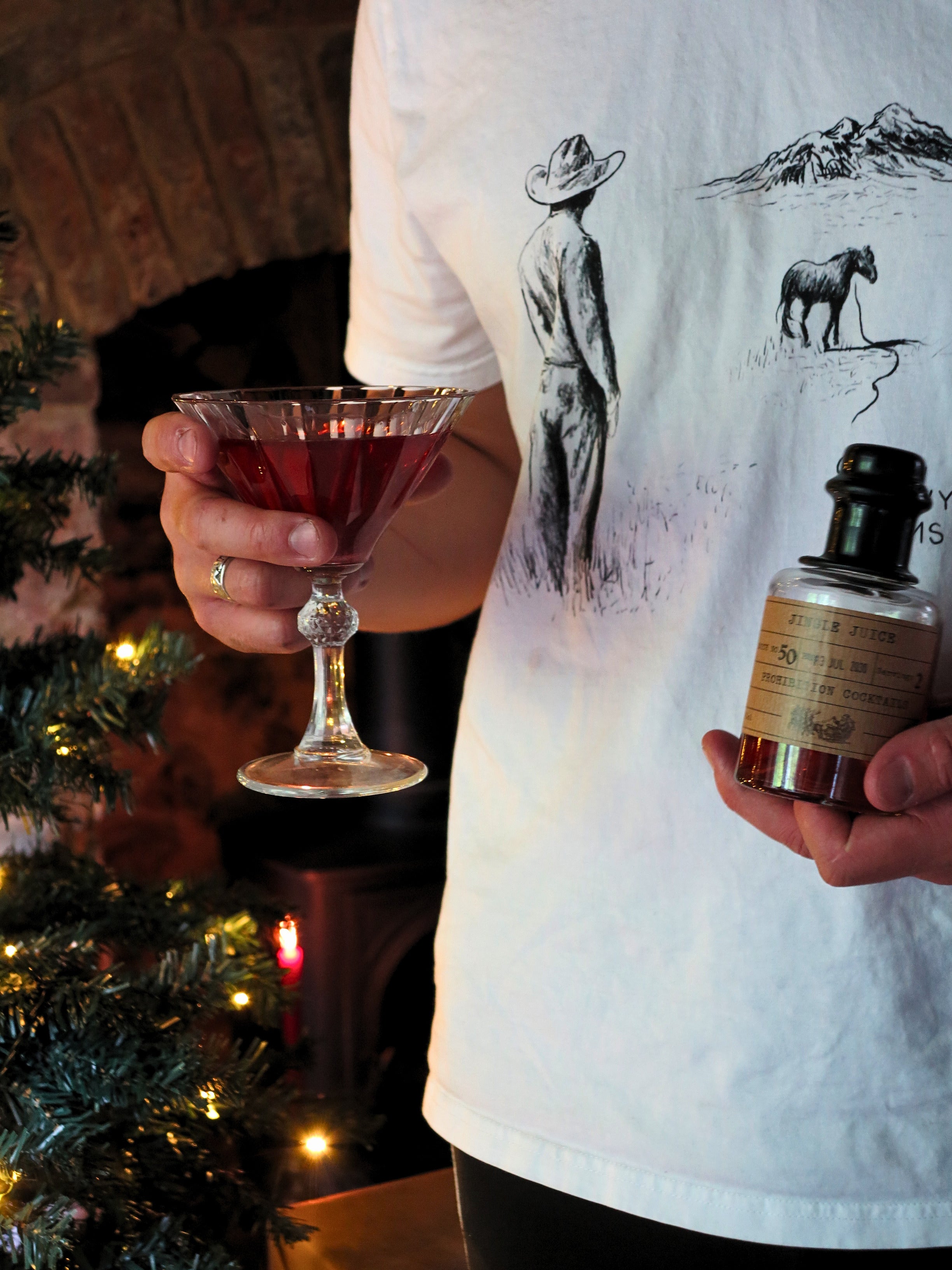 A person in a white graphic t-shirt holds a small bottle in one hand Jingle Juice by Prohibition Cocktails and a martini glass with red drink in the other, posed in front of a fireplace and a Christmas tree.