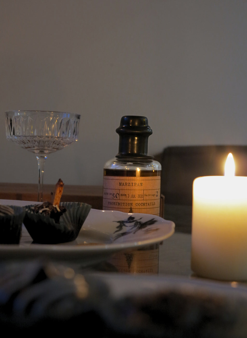 A lit candle and a small bottle labeled 'MARZIPAN' sit on a table beside a plate with dark cupcakes in a softly lit indoor setting.