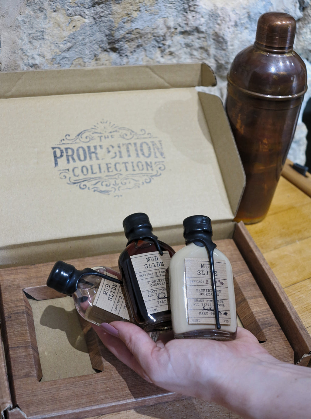 A hand holds three small bottles from The Prohibition Collection inside a wooden box, with a copper-colored cocktail shaker in the background against a stone wall.