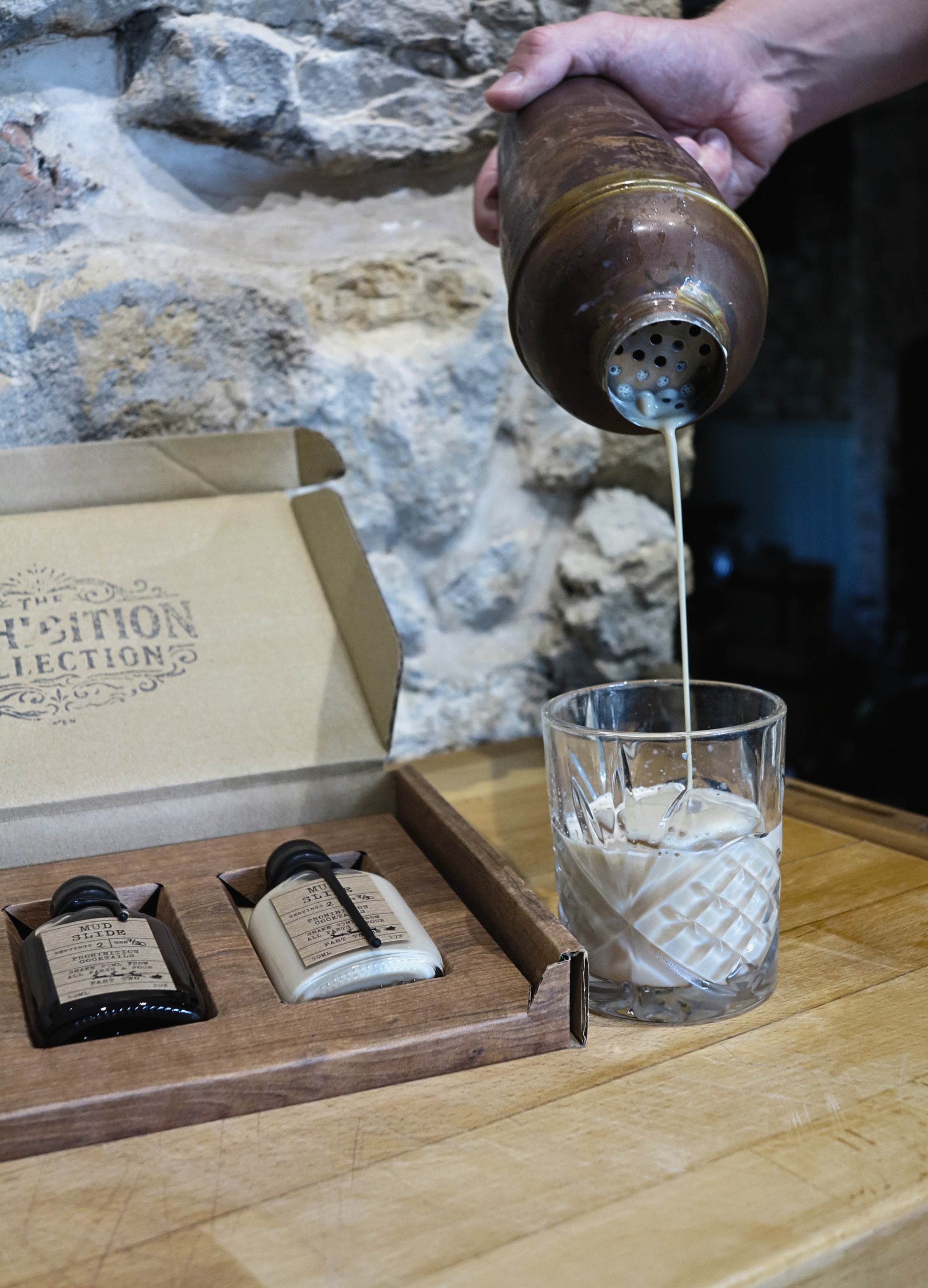 A hand pours a drink from a metal shaker into a clear textured glass on a wooden table, with a branded box and two small bottles in the background against a stone wall.