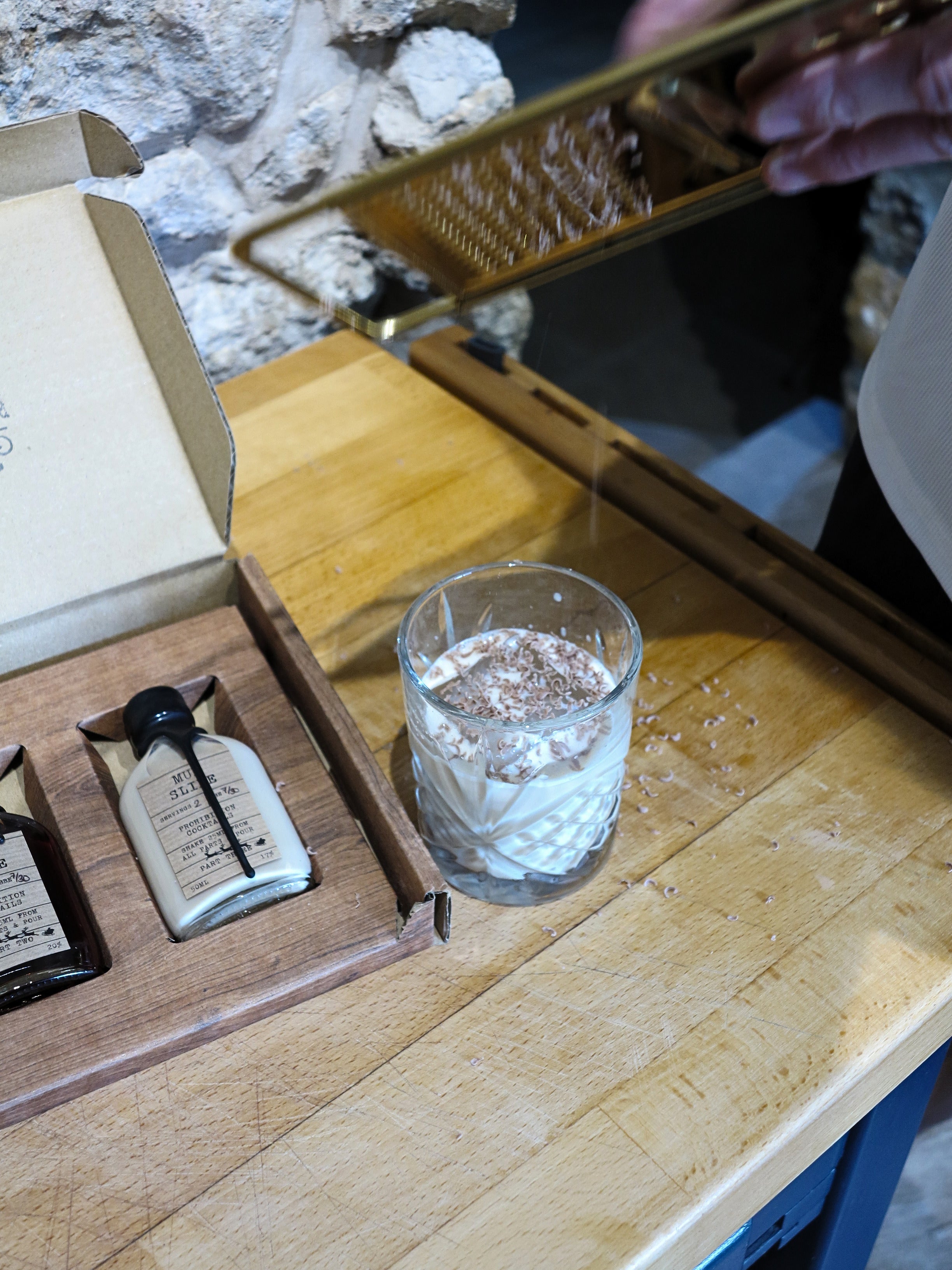 A small clear glass tumbler filled with a white beverage and topped with cocoa powder, set on a wooden table next to an open gift box containing two cream-colored bottles. A hand is holding a gold grater above the glass.