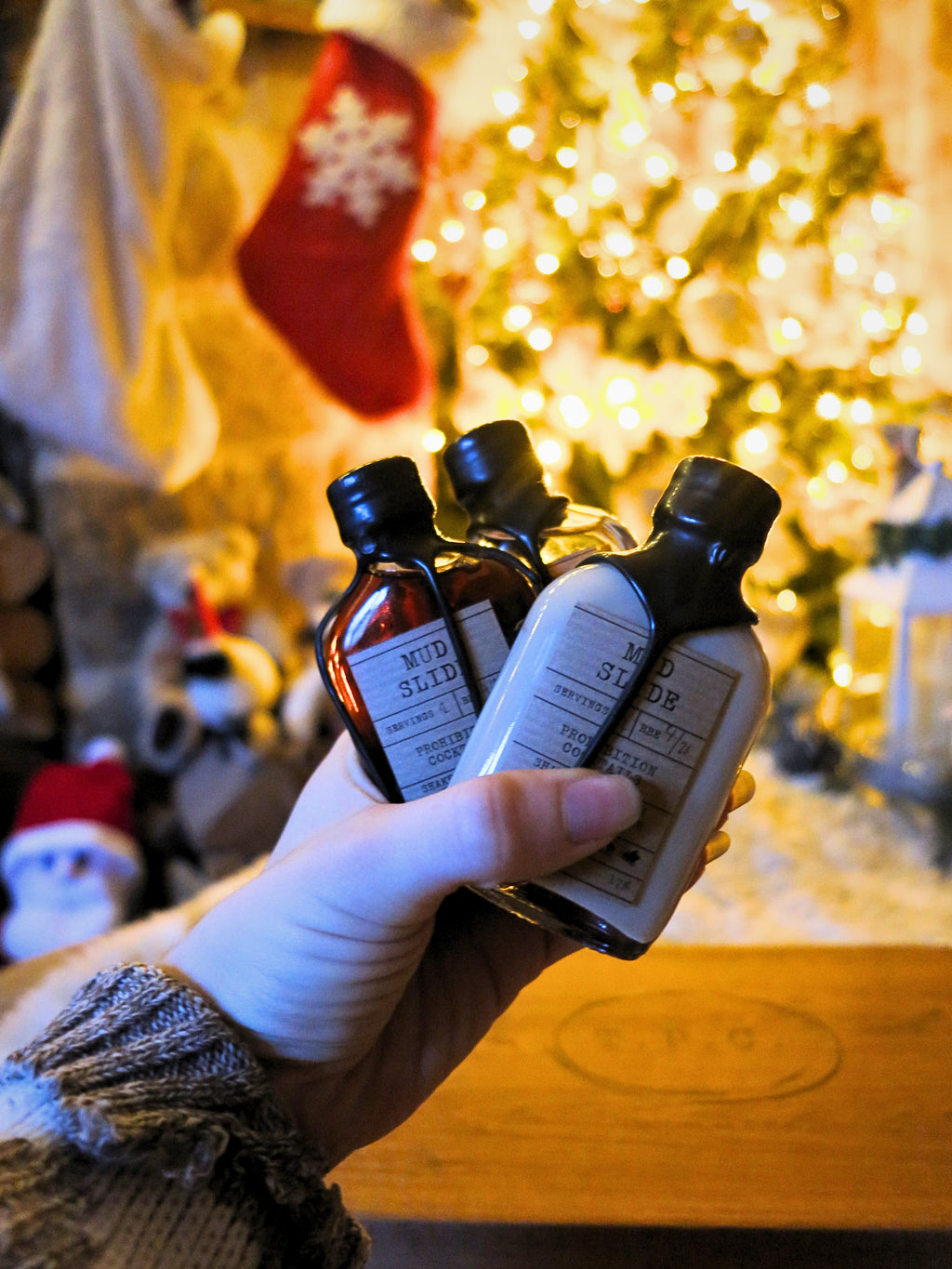 A hand holds three bottles in a cozy, warmly lit holiday setting with a blurred Christmas tree and stockings in the background. The bottles have black caps and white labels, with visible text including 'MUD SLIDE' 'Prohibition Cocktails'