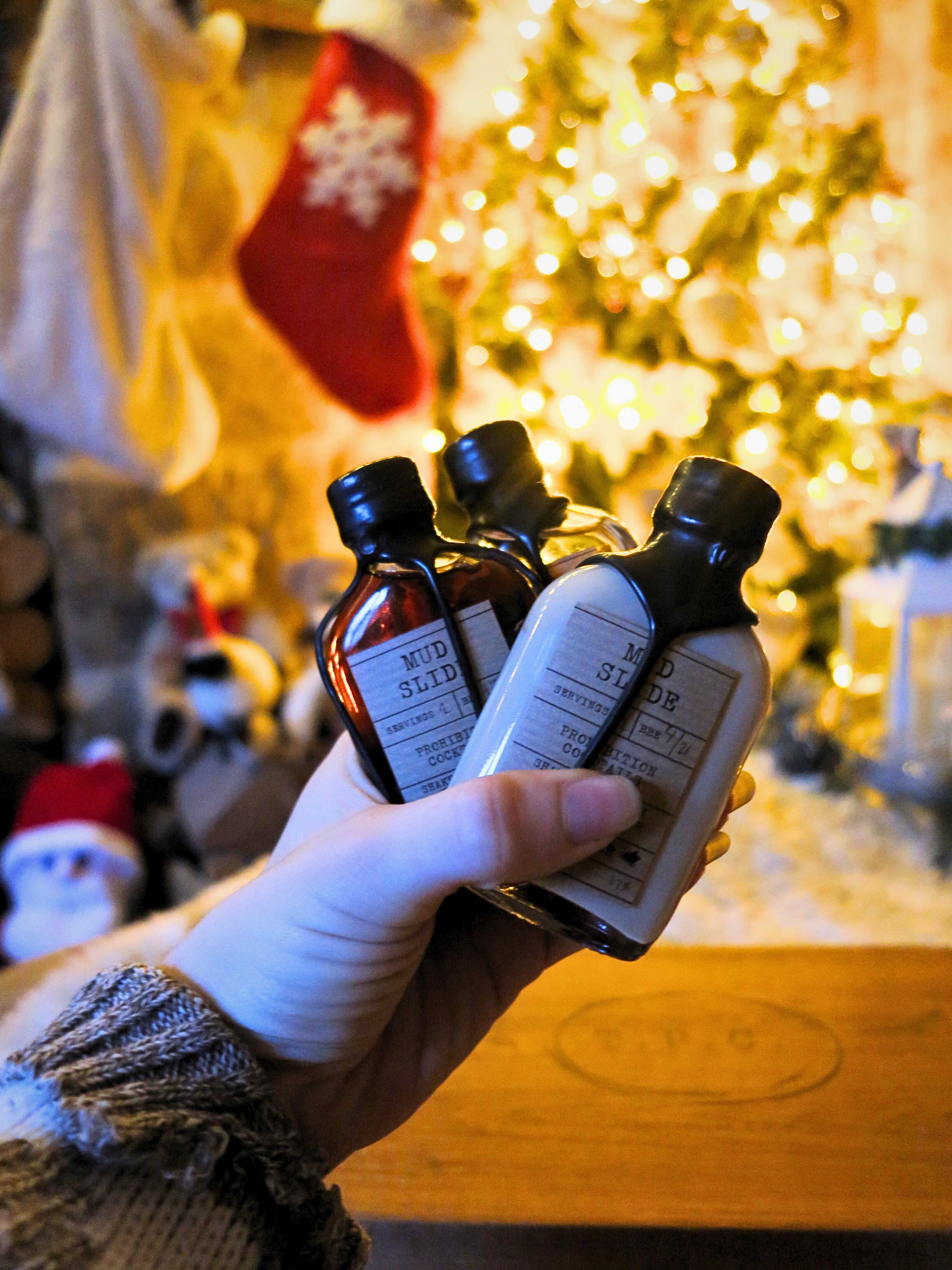A hand holds three bottles in a cozy, warmly lit holiday setting with a blurred Christmas tree and stockings in the background. The bottles have black caps and white labels, with visible text including 'MUD SLIDE' 'Prohibition Cocktails'