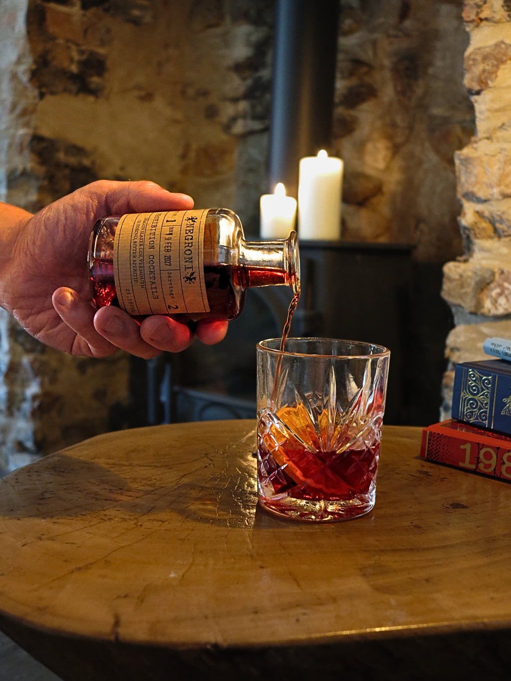 A hand pours a dark amber liquid - Negroni by Prohibition Cocktails - from a small bottle with a cream label into a cut-glass tumbler on a rustic wooden table, with candles and stone walls in the background.