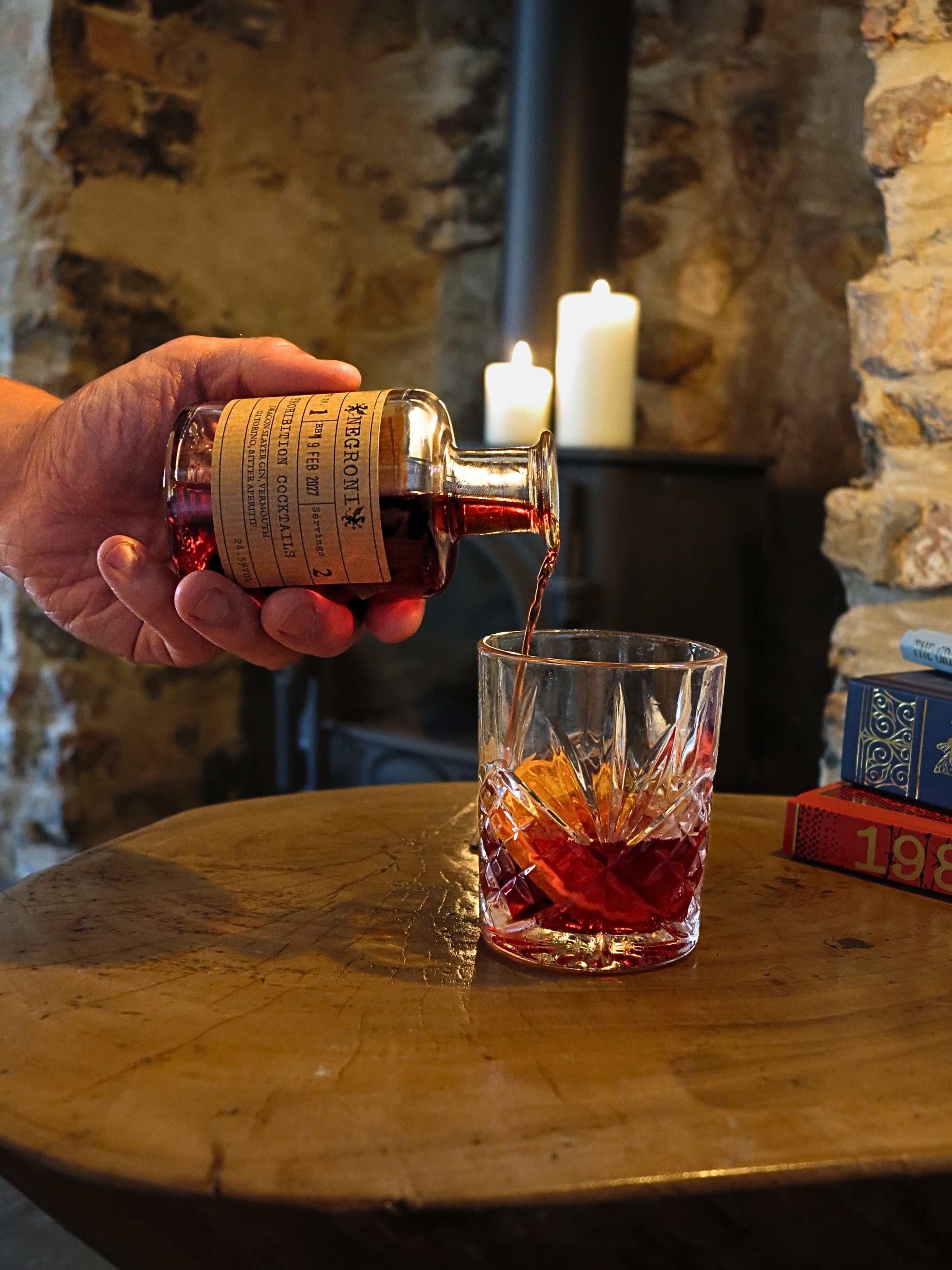 A hand pours a dark amber liquid - Negroni by Prohibition Cocktails - from a small bottle with a cream label into a cut-glass tumbler on a rustic wooden table, with candles and stone walls in the background.