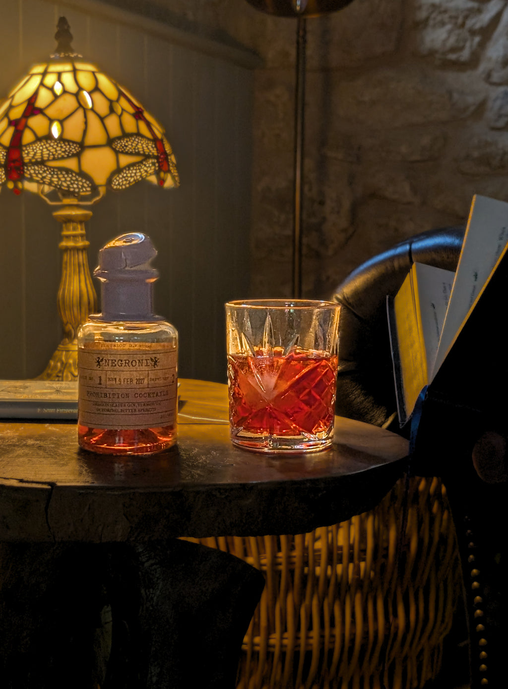 A bottle of whiskey and a cut-glass tumbler filled with amber liquor - Negroni by Prohibition Cocktails - sit on a dark wooden table in a dimly lit room with a stained-glass lamp, stone wall, and open book.