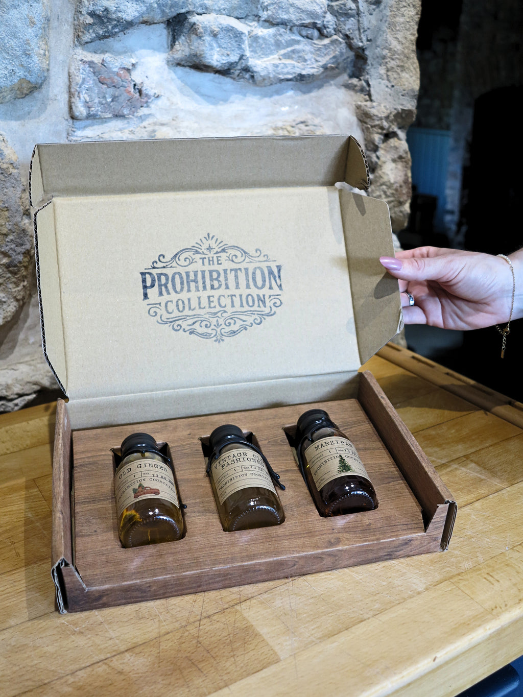 A hand opens a cardboard box labeled 'The Prohibition Collection' resting on a wooden table, revealing three small amber glass bottles in a wooden tray.