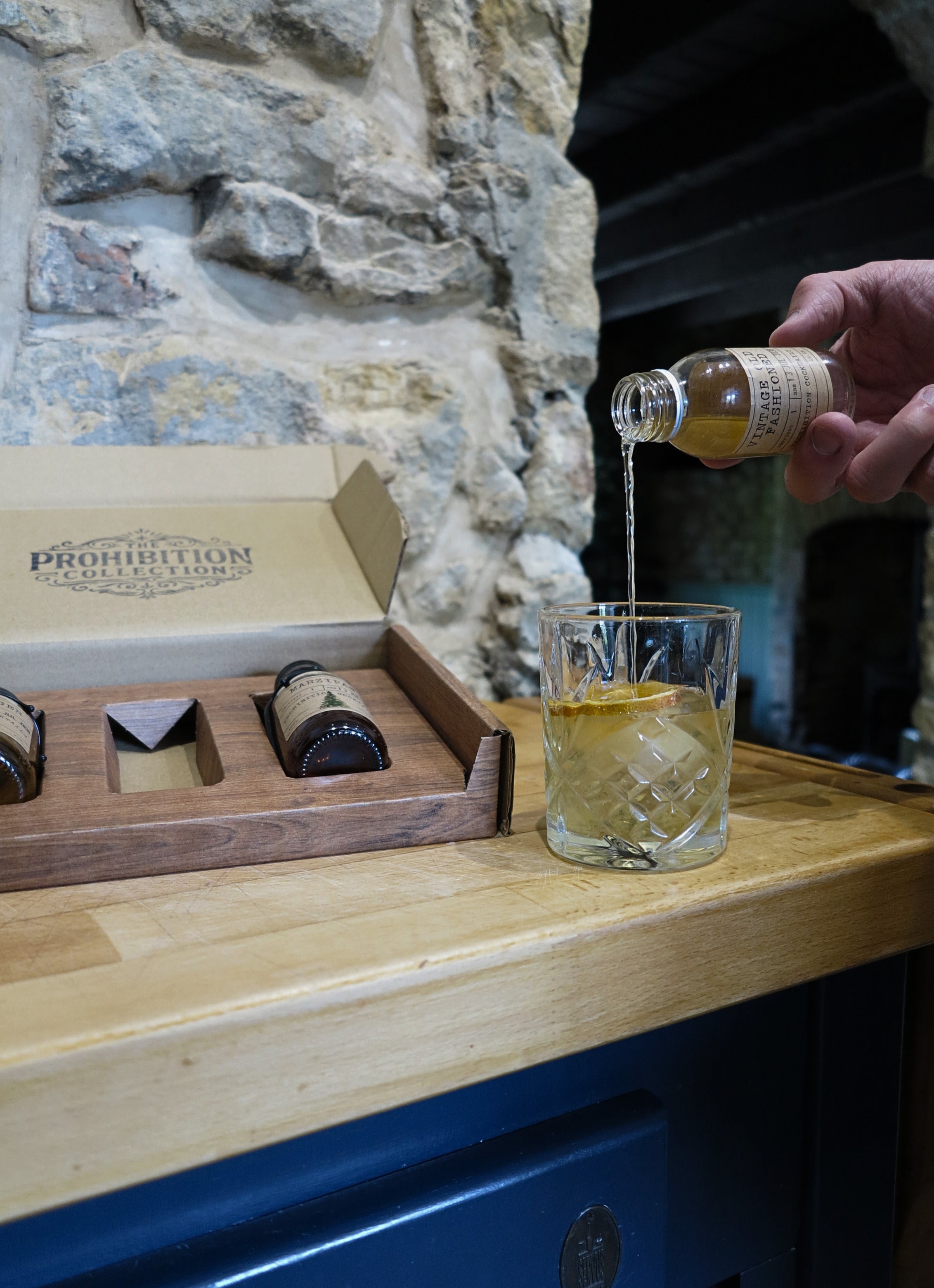 A hand pours a clear liquid from a small bottle into a cut-glass tumbler on a wooden counter, with a branded Prohibition Collection gift box and several small bottles in the background against a stone wall.