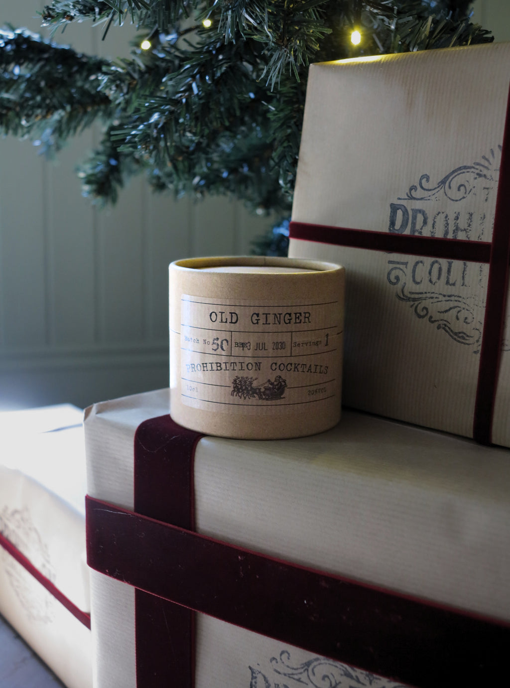 A cylindrical candle jar labeled 'OLD GINGER' sits on top of wrapped gift boxes with dark red ribbon, with a Christmas tree branch and warm string lights in the background.