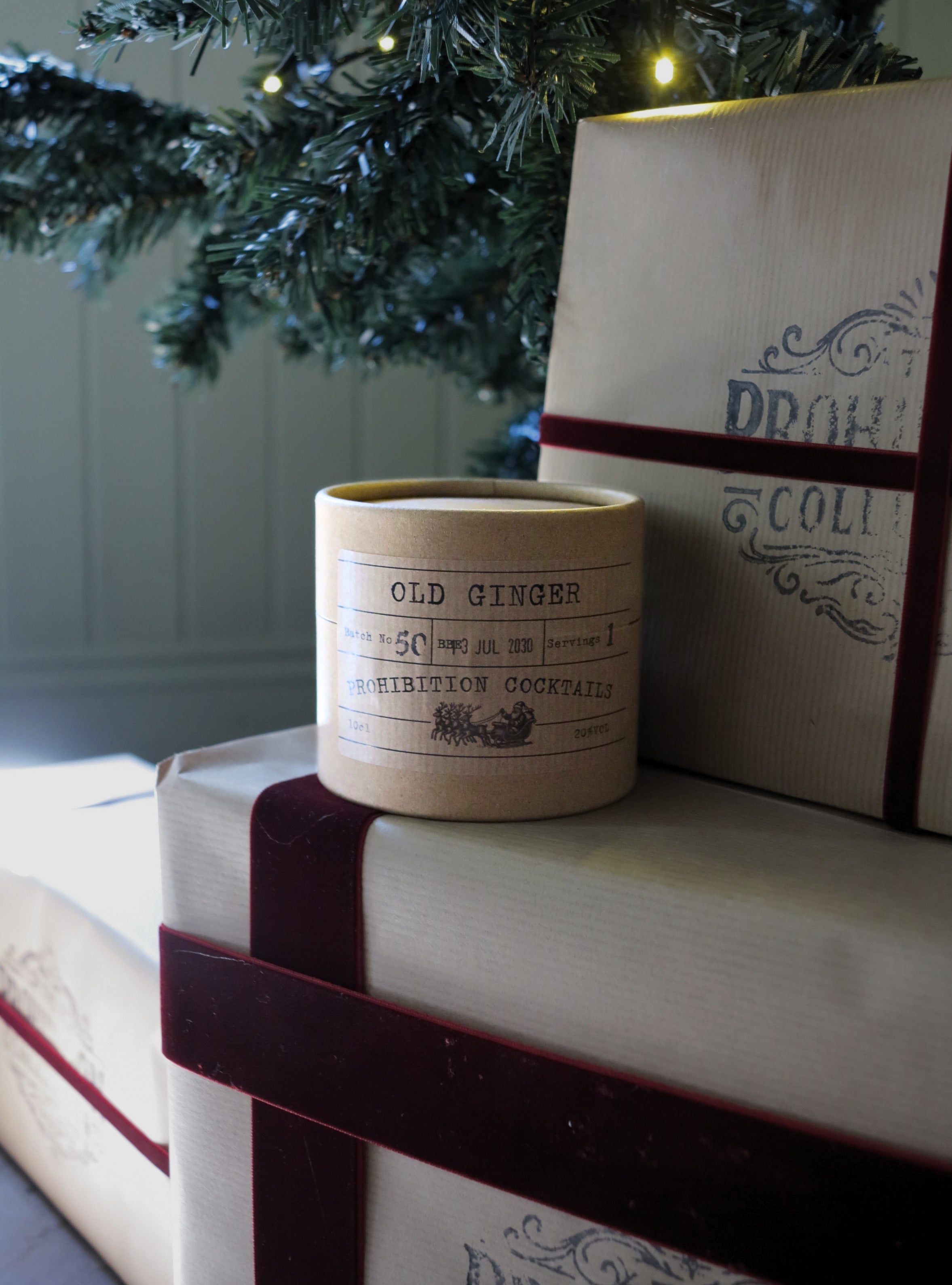 A cylindrical candle jar labeled 'OLD GINGER' sits on top of wrapped gift boxes with dark red ribbon, with a Christmas tree branch and warm string lights in the background.