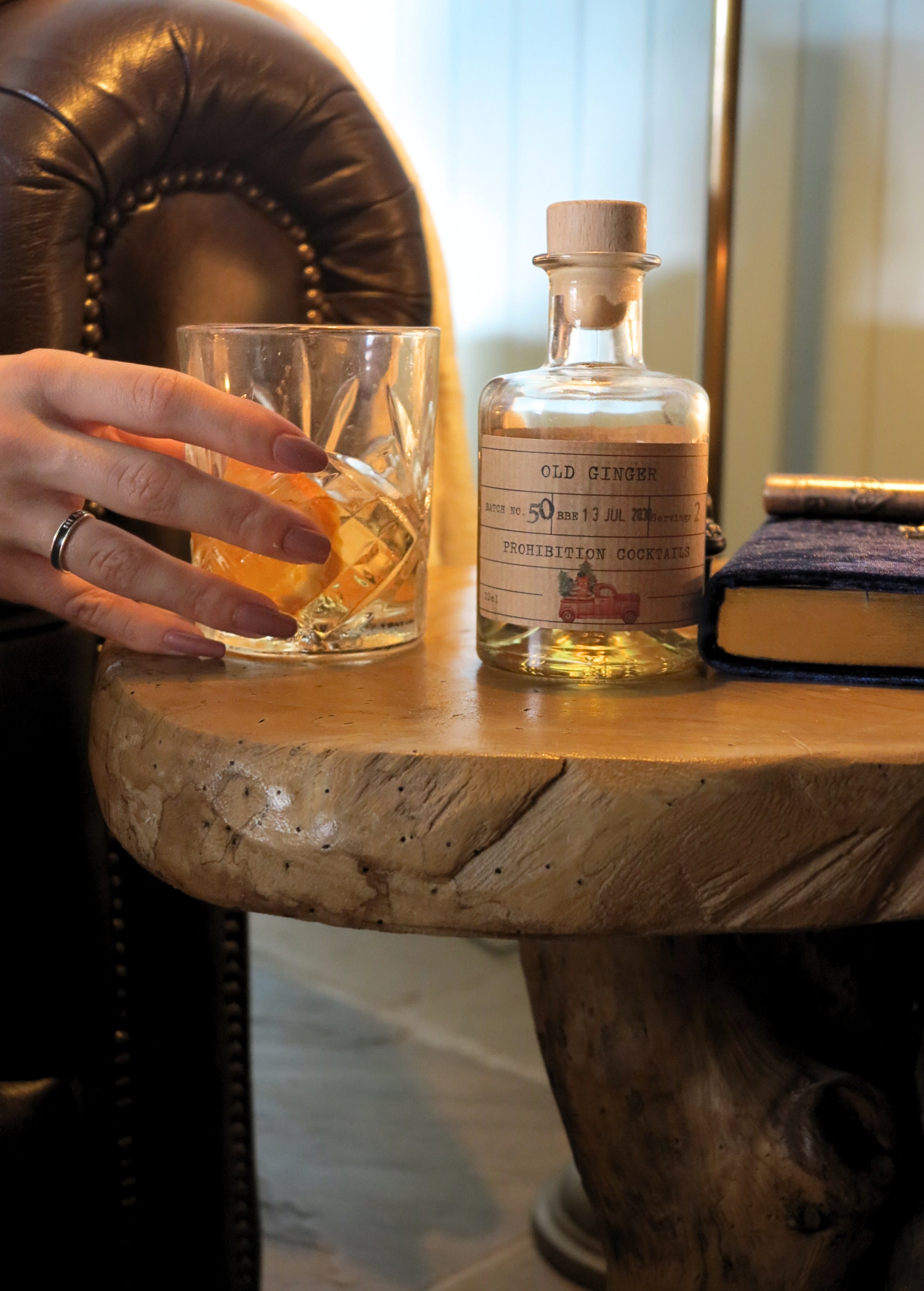 A hand holding a cut-glass tumbler filled with amber liquid beside a small clear bottle labeled 'OLD Ginger' on a rustic wooden table, with a leather chair and books in the background.