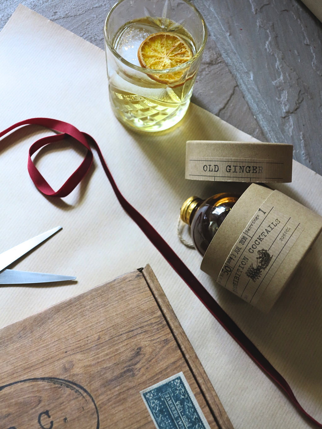 A styled flat-lay of a bottle of Old Ginger by Prohibition Cocktails with kraft paper labels, a glass of the golden gin with a dried orange slice, scissors, a wooden box, and a red ribbon on a neutral background.