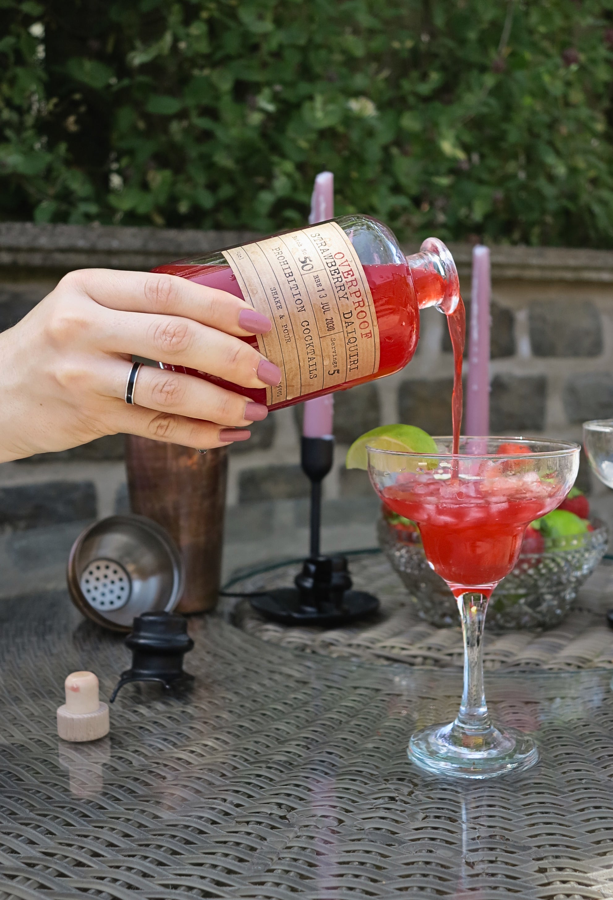 A hand pours an overproof strawberry daiquiri from Prohibition Cocktails from a bottle with a tan paper label into a coupe glass on an outdoor patio table.
