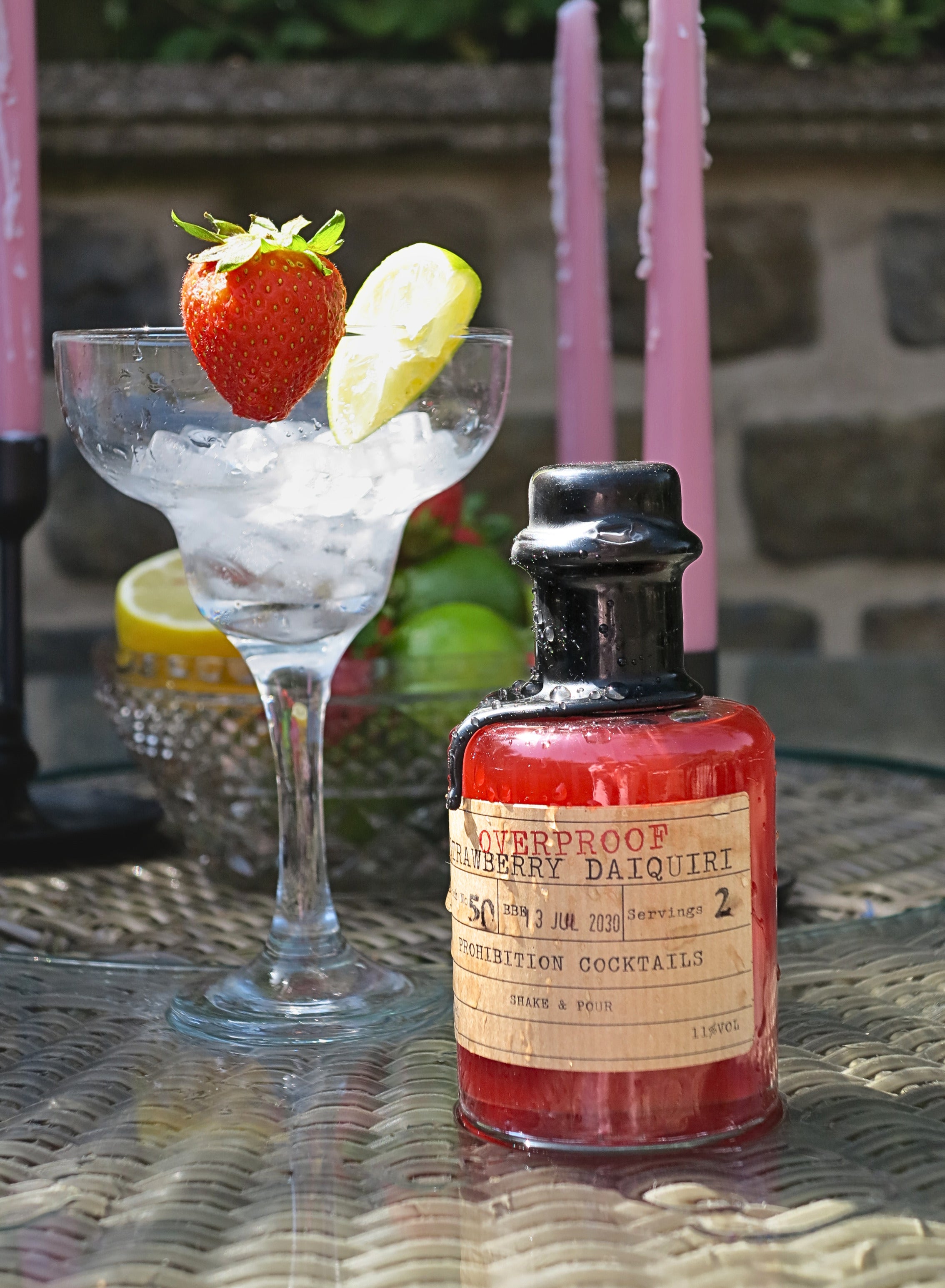 A small bottle of Overproof Strawberry Daiquiri cocktail mix from Prohibition Cocktails stands on a woven table next to a margarita glass filled with ice, garnished with a strawberry and lime wedge, with pink candles in the background.