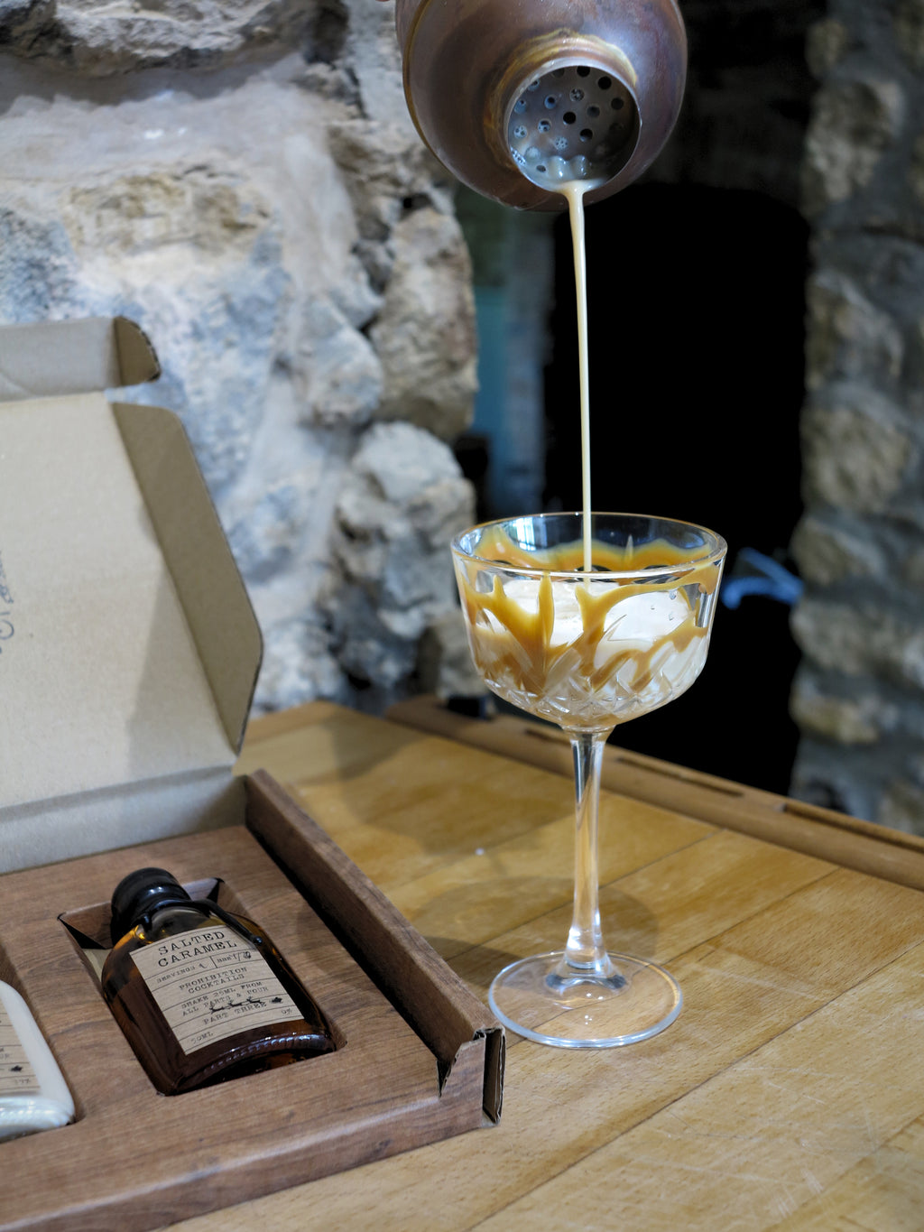 A small bottle labeled Salted Caramel is shown in an open presentation box on a wooden table, with a cocktail shaker pouring a pale drink into a coupe glass nearby. The background features a rustic stone wall.