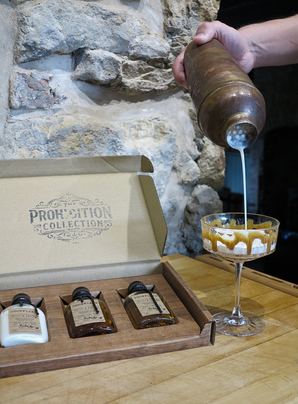 A hand pours a creamy white liquid from a metal shaker into a coupe glass with caramel drizzle, with an open box labeled 'The Prohibition Collection' containing small bottles on a wooden table against a stone wall.