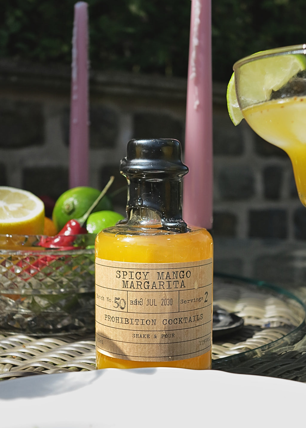 A bottle of 'Spicy Mango Margarita' prohibition cocktail mix, filled with bright orange liquid and featuring a vintage-style paper label, set on a woven table with a margarita glass and fresh citrus in the background.
