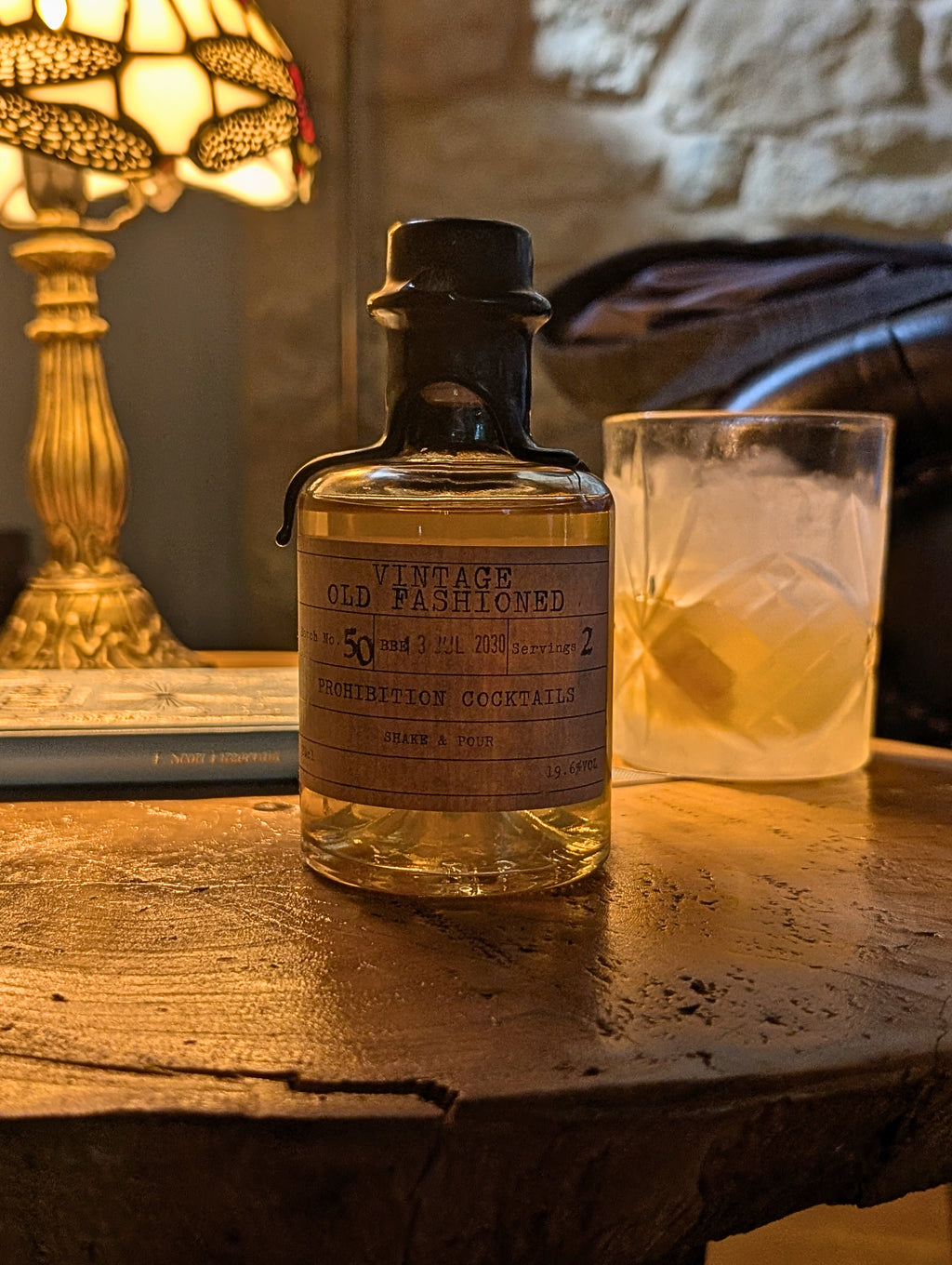 A bottle of Martini cocktail mix labeled 'Vintage Old Fashioned' sits on a rustic wooden table next to a glass of cocktail, with a warm lamp and stone wall in the background.