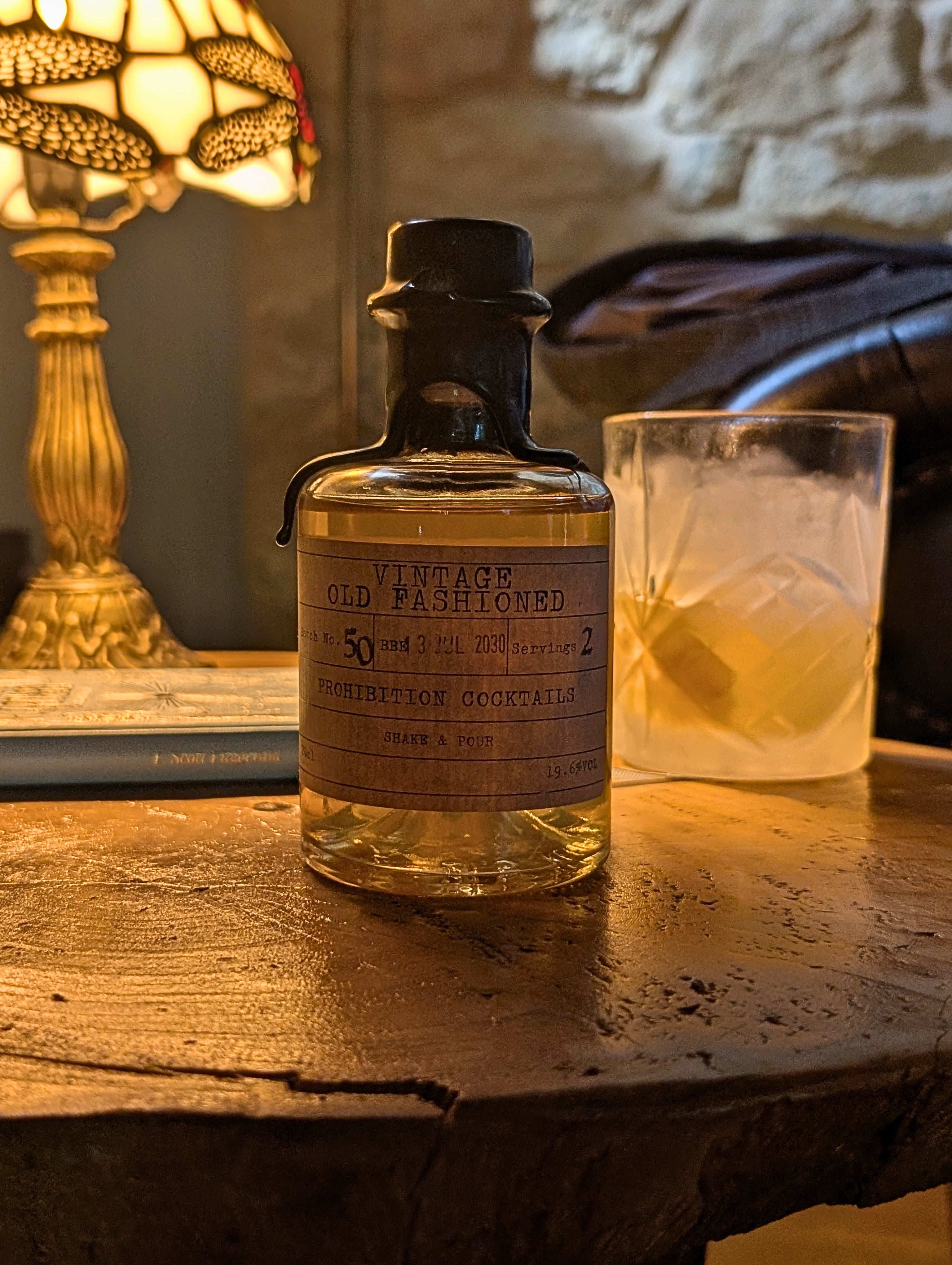 A bottle of Martini cocktail mix labeled 'Vintage Old Fashioned' sits on a rustic wooden table next to a glass of cocktail, with a warm lamp and stone wall in the background.