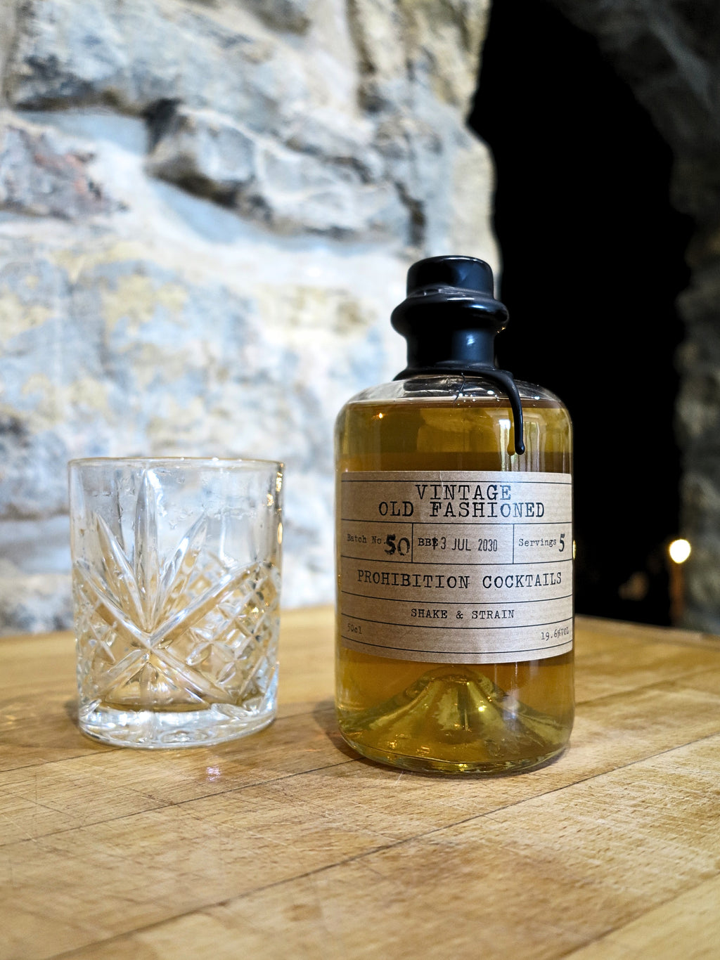 A bottle of 'Vintage Old Fashioned' prohibition cocktails with amber liquid, next to a cut-crystal tumbler, set on a wooden table with a rustic stone wall background.