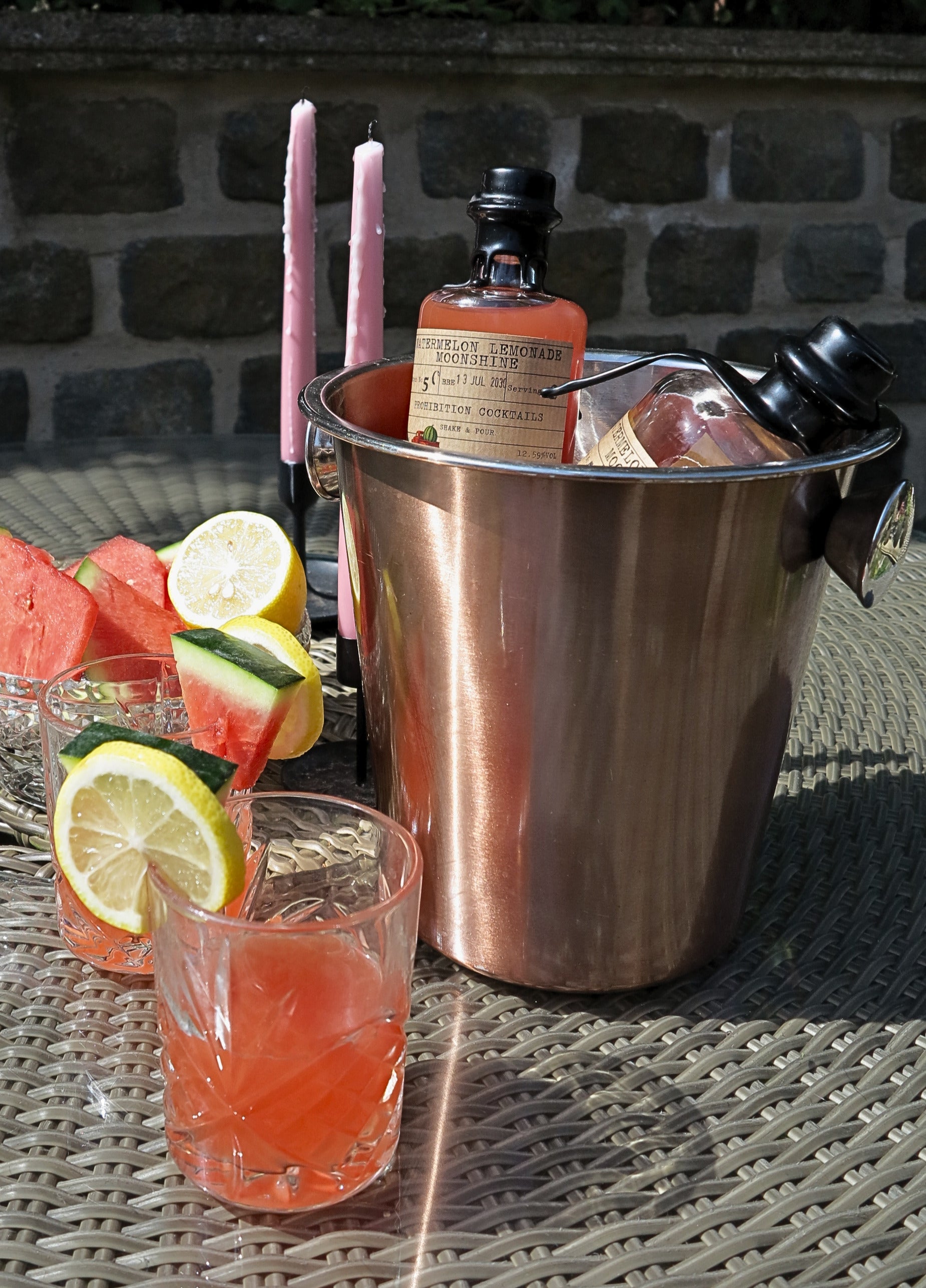 A copper cocktail bucket filled with ice and pink drinks, set on an outdoor patio table with two glasses of pink beverage garnished with watermelon and lemon slices, pink taper candles, and a bottle in the background.