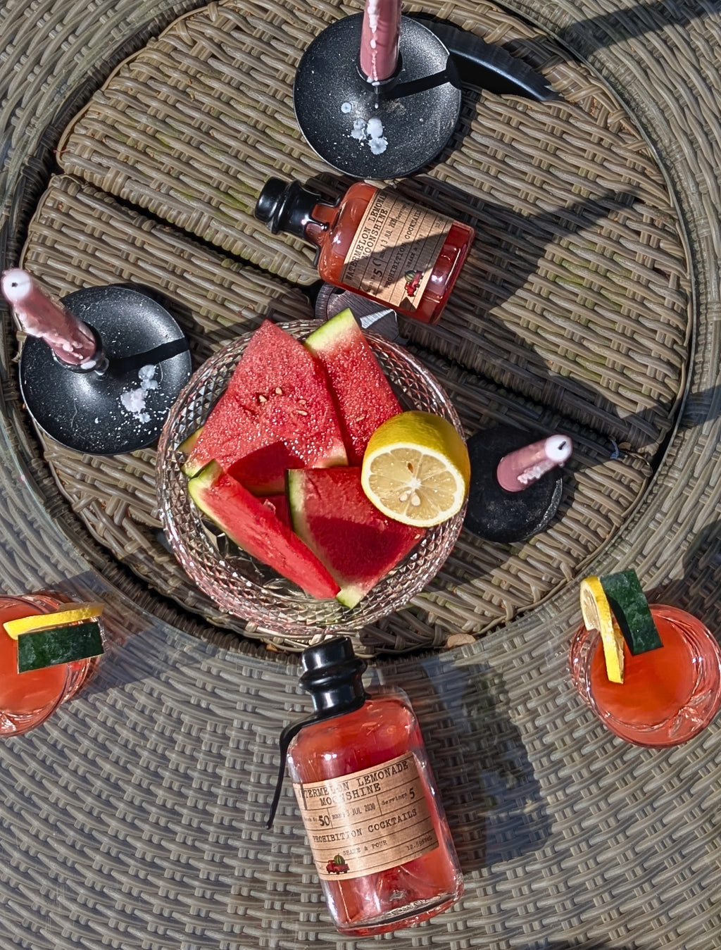 A styled overhead flat-lay of a cocktail-making setup on a woven round table. A glass bowl holds watermelon slices and a lemon wedge, surrounded by several bottles of pink-red liquids and garnished drinks with cucumber and lemon slices.